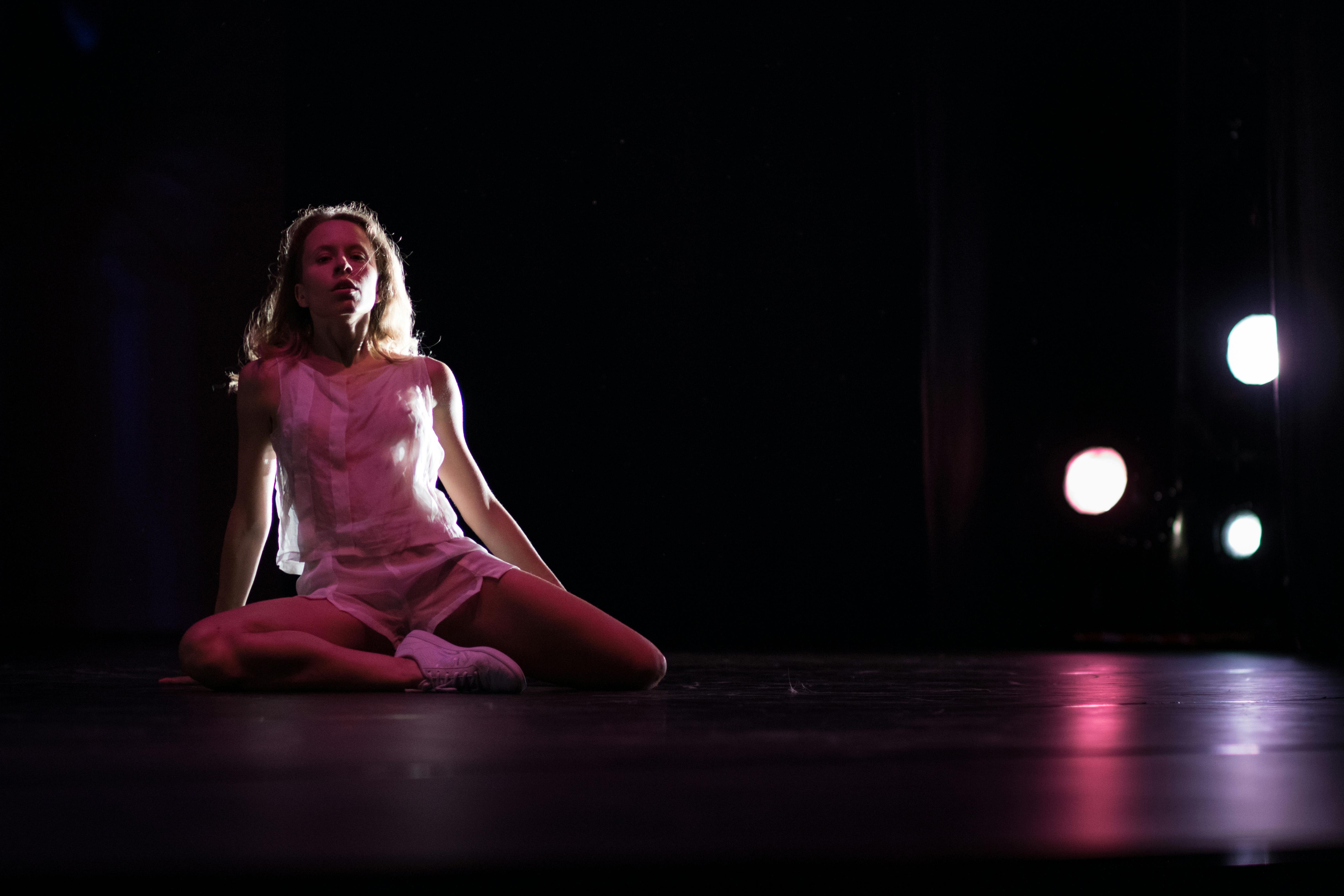 Una ballerina in scena, indossa vestito e sneakers bianche. è seduta a terra, ha le gambe incrociate e guarda in alto.