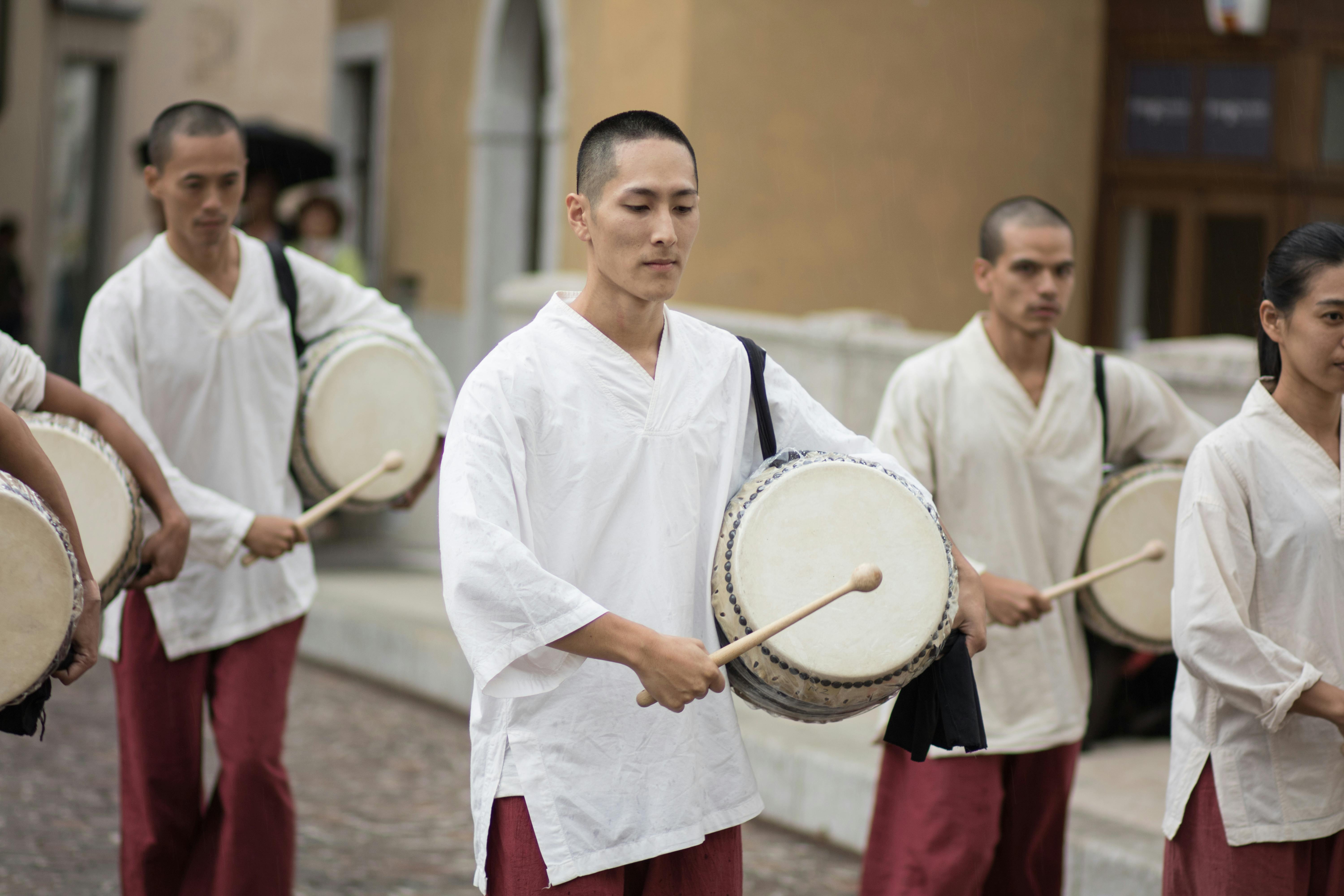 Alcuni artisti in strada. Camminano in parata con dei tamburi sotto al braccio. Indossano camicie bianche e pantaloni bordeaux