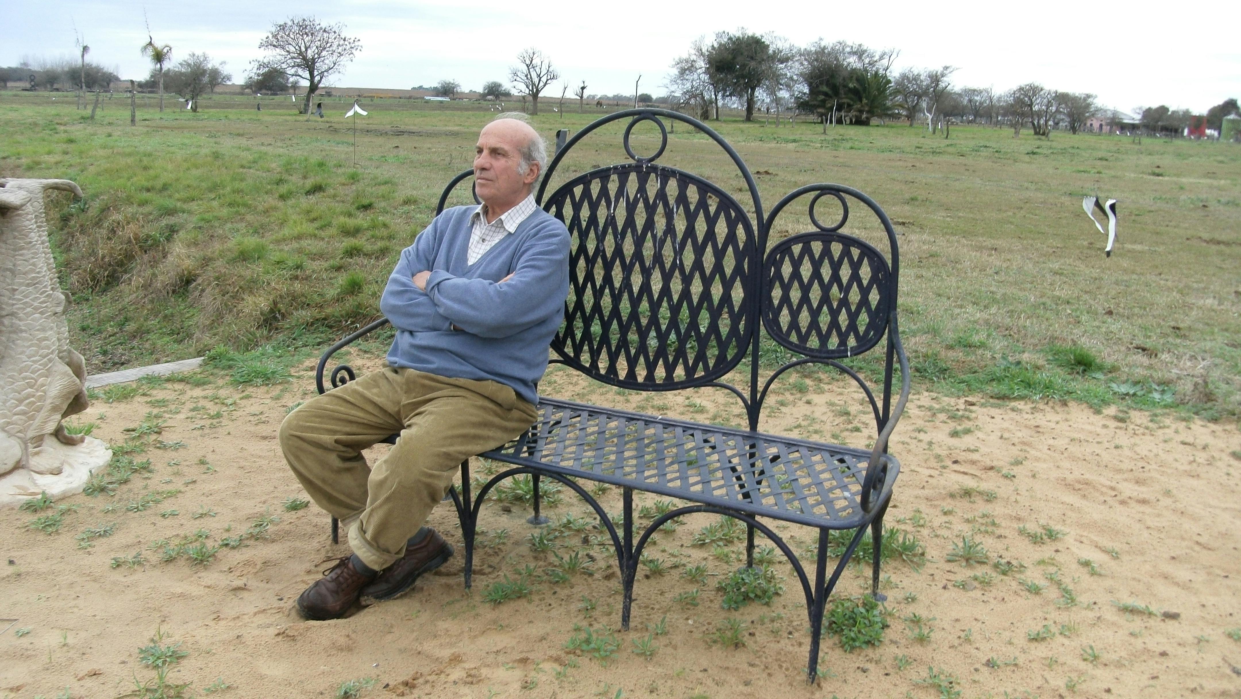 In the photo, Enrico Pugliese is sitting with his arms crossed on a bench in the middle of a field.