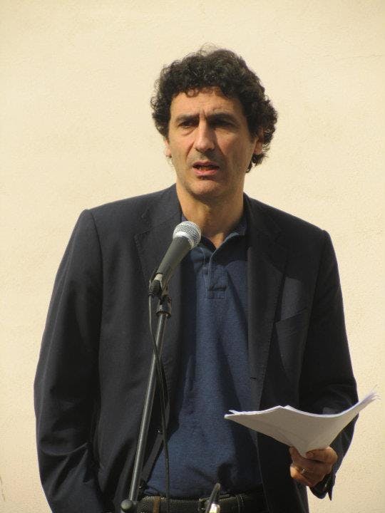 In the photo, Paolo Borioni is speaking into a microphone while holding some papers in his hand. He is standing, wearing a black jacket and a blue polo shirt.