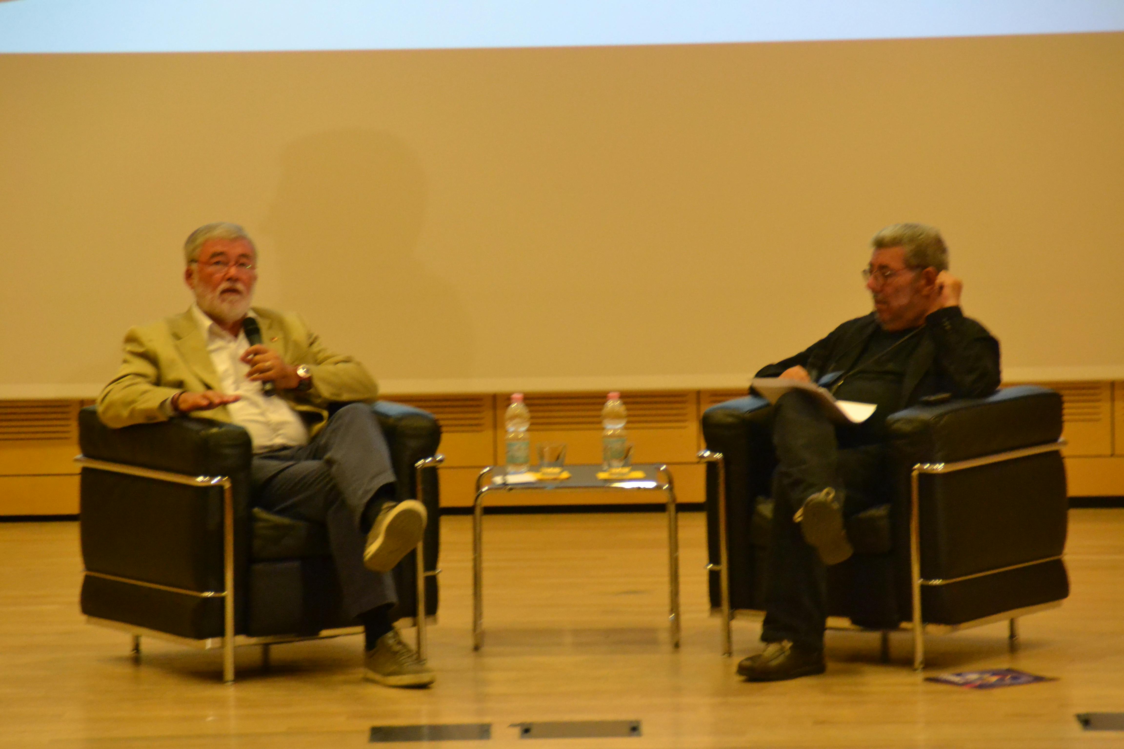 Foto di Sergio Cofferati, parlamentare europeo e Mario Cossali, vicesindaco di Rovereto e Isera, mentre parlano seduti su due sedie sul palco.