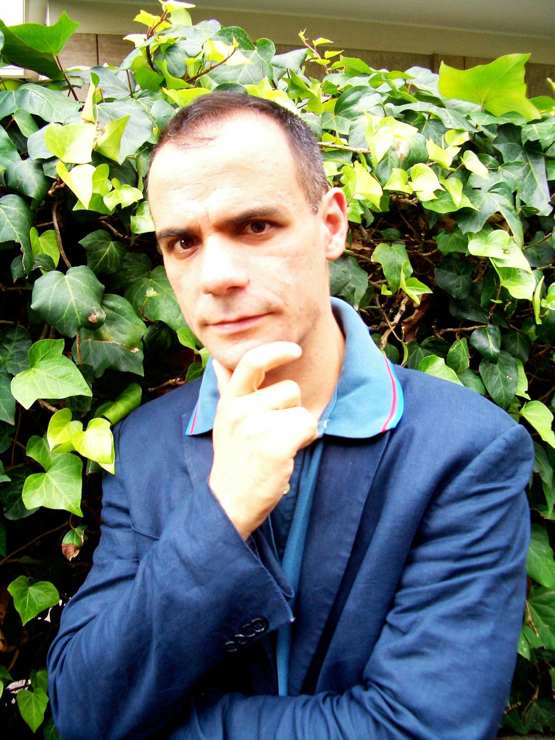 Ritratto di Teodoro Andreadis Synghellakist. Ha le braccia conserte e la mano destra appoggiata al proprio mento. Indossa polo azzurra e giacca blu.
