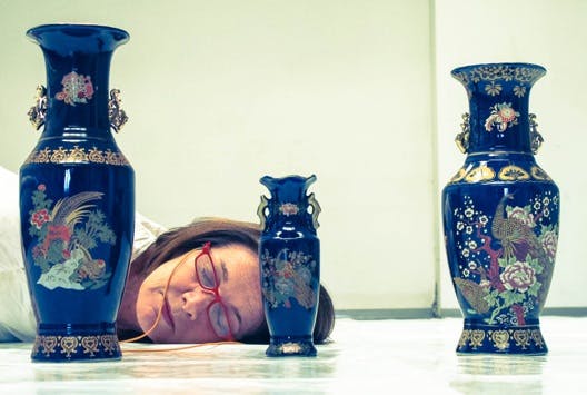 In the foreground, three blue vases resting on the floor. Behind them, the face of a woman lying down with her eyes closed.