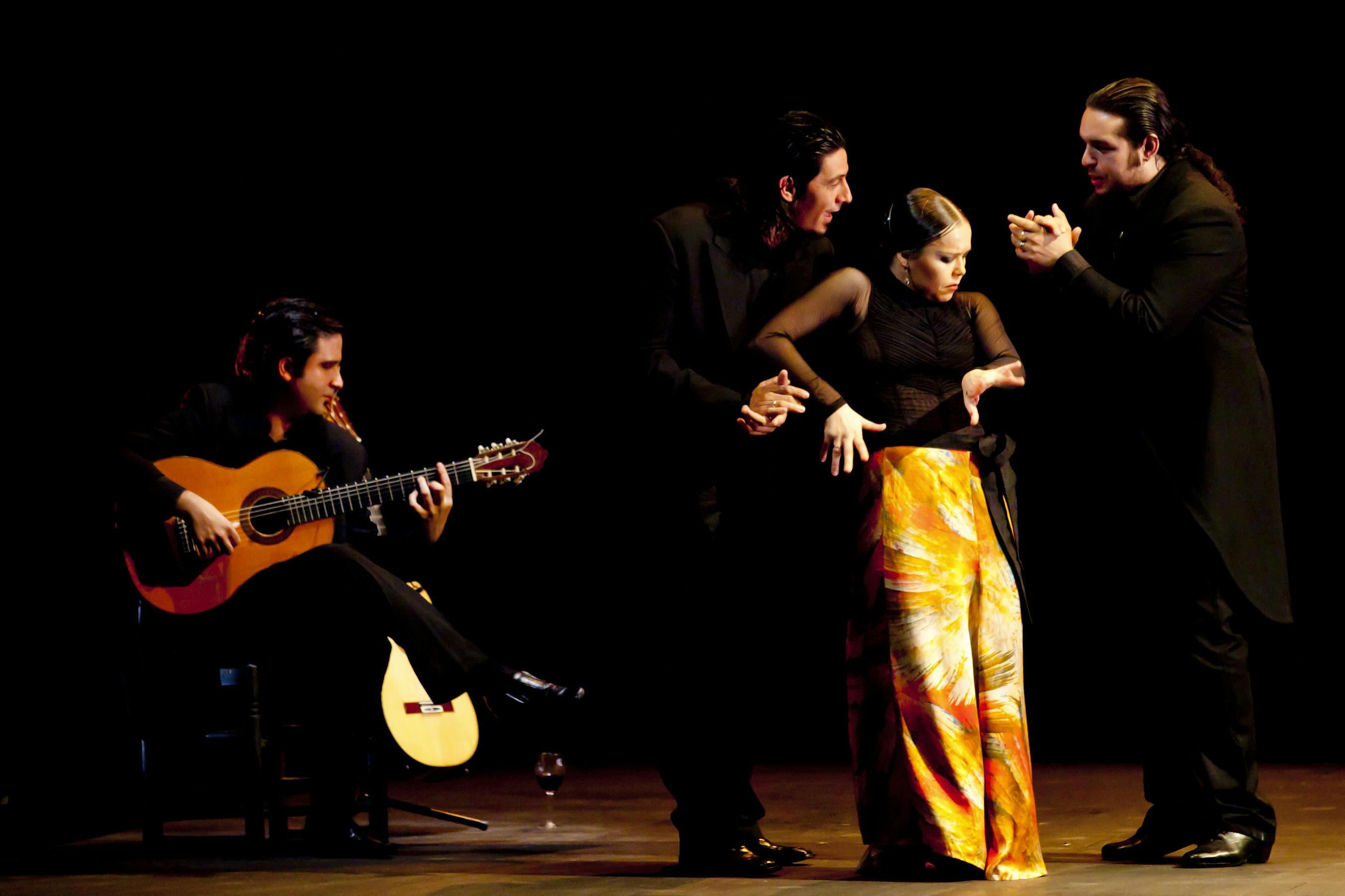 Three dancers on stage, standing. Two men are wearing black suits, and the female dancer is in yellow pants. Behind them, there is a guitarist.