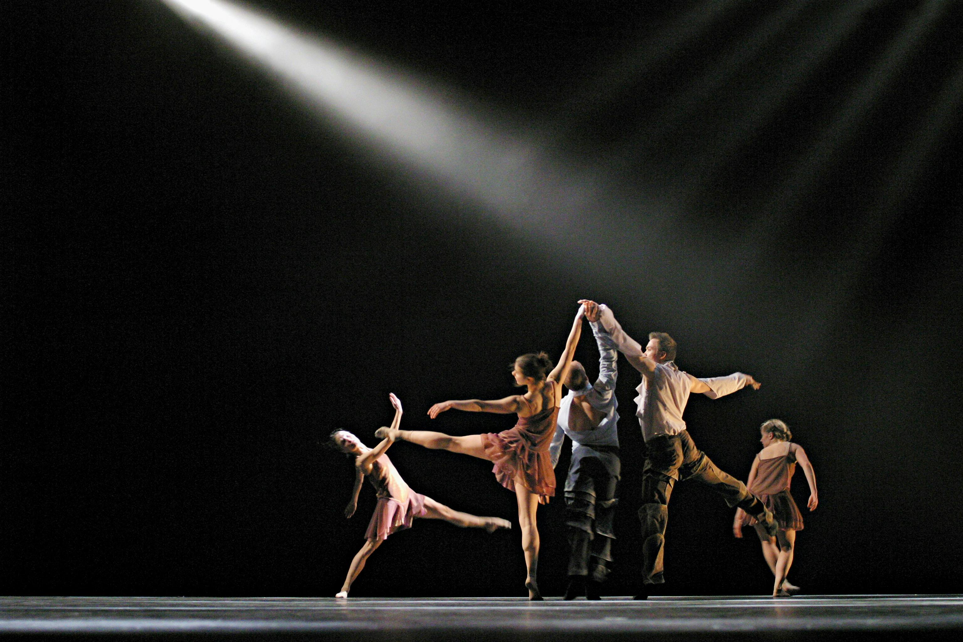 Five dancers on stage. They are standing close together, each in different poses. A beam of light shines on them from the left.