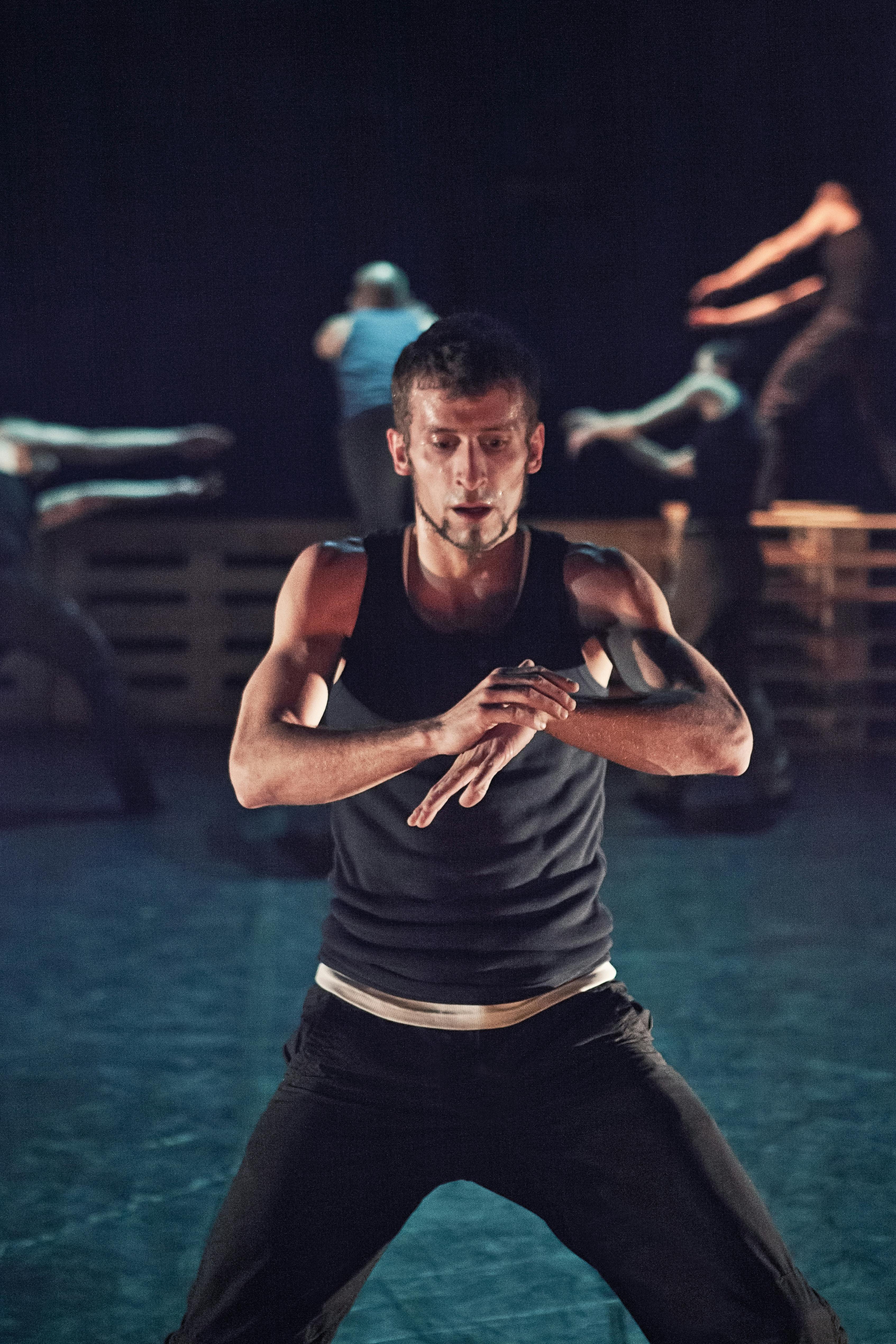 In the foreground, a dancer holds his hands crossed in front of him. Behind him, other artists are on pallets.