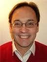 In the photo, Giovanni Scotto is smiling at the camera. He is wearing glasses, a white shirt, and a red sweater.