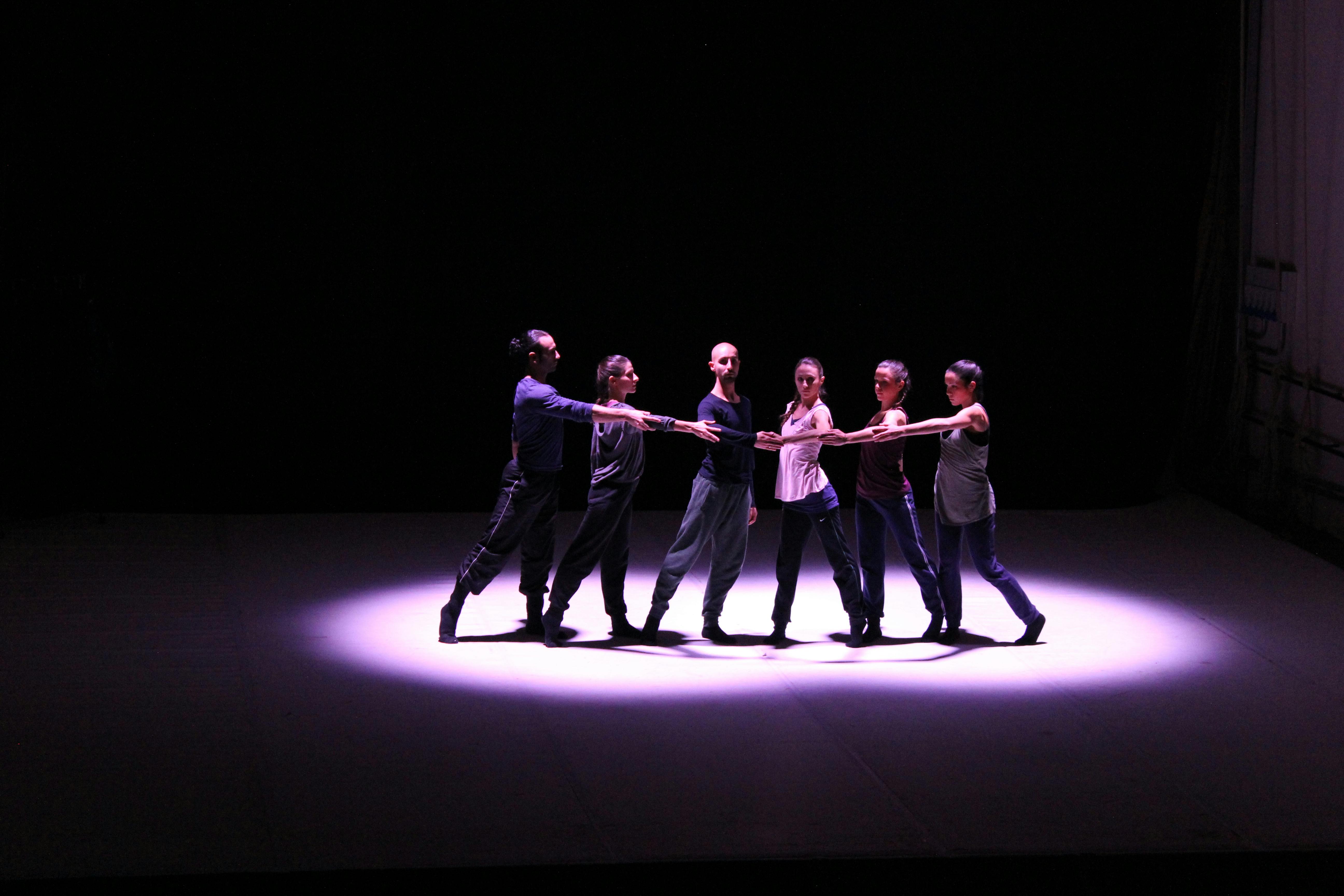Six artists stand with one leg extended backward, reaching one arm toward the same point. They are illuminated from the side by a spotlight.