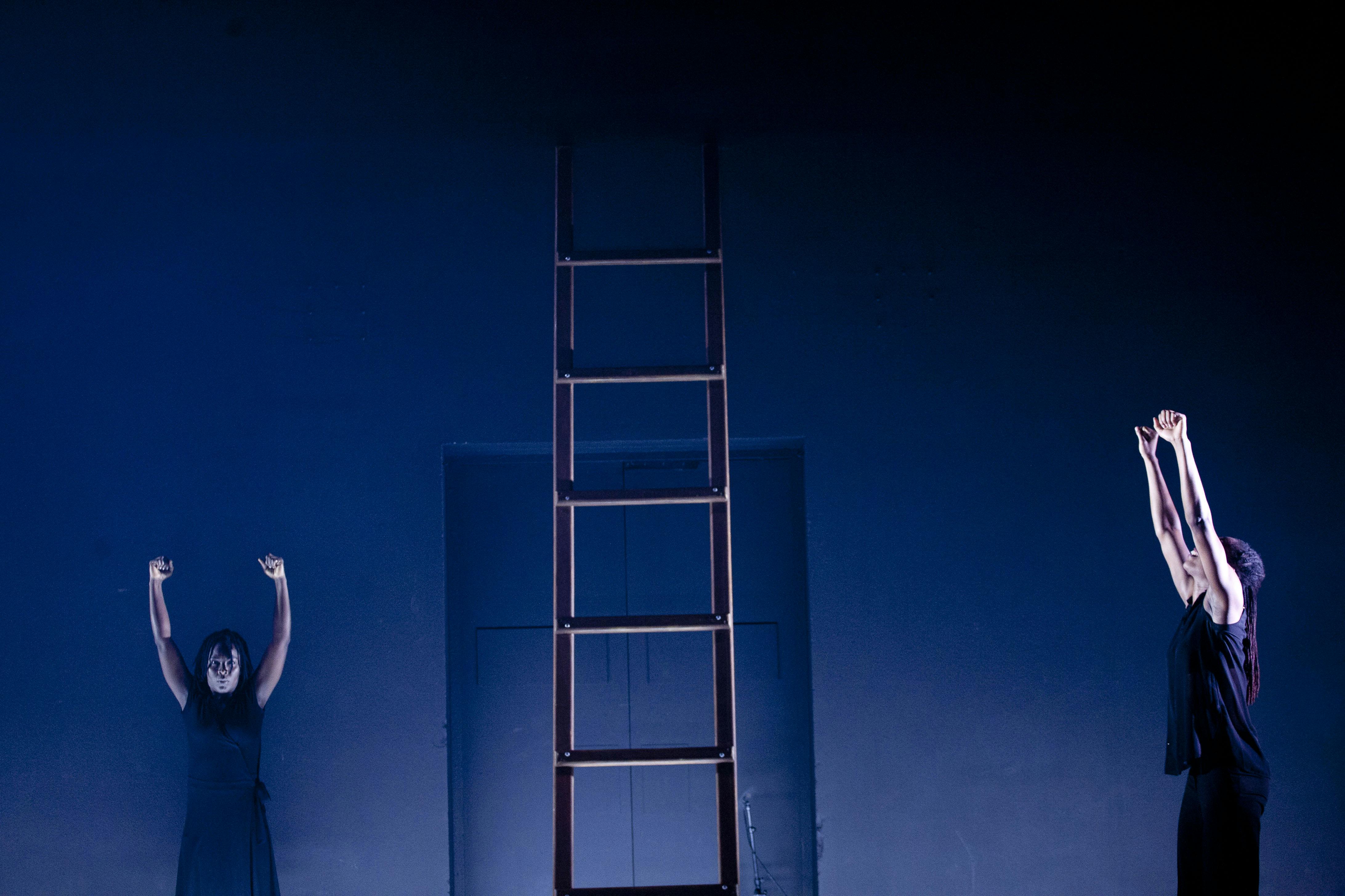 Due artisti in scena. Entrambi hanno le braccia tese in alto. In mezzo alla scena una scala a pioli