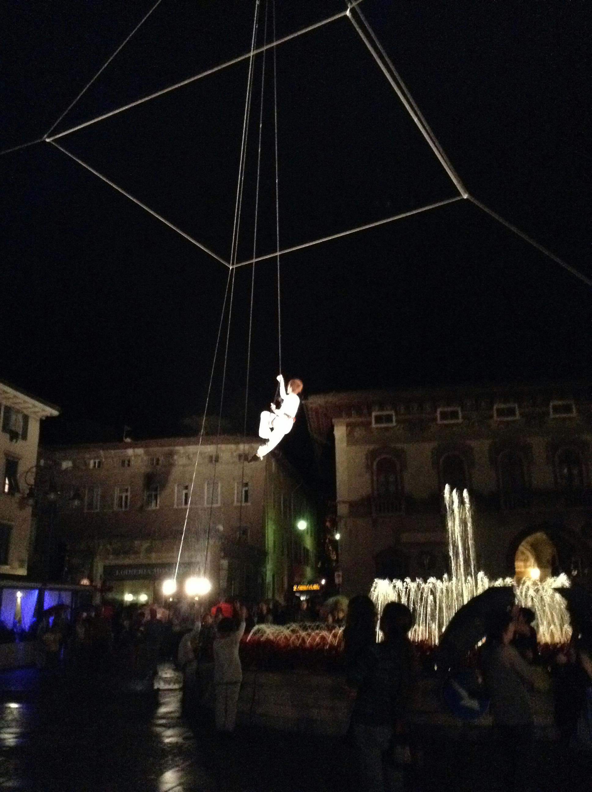 In piazza a Rovereto, di sera, degli artisti sono sospesi a mezz'aria su un'impalcatura quadrata.