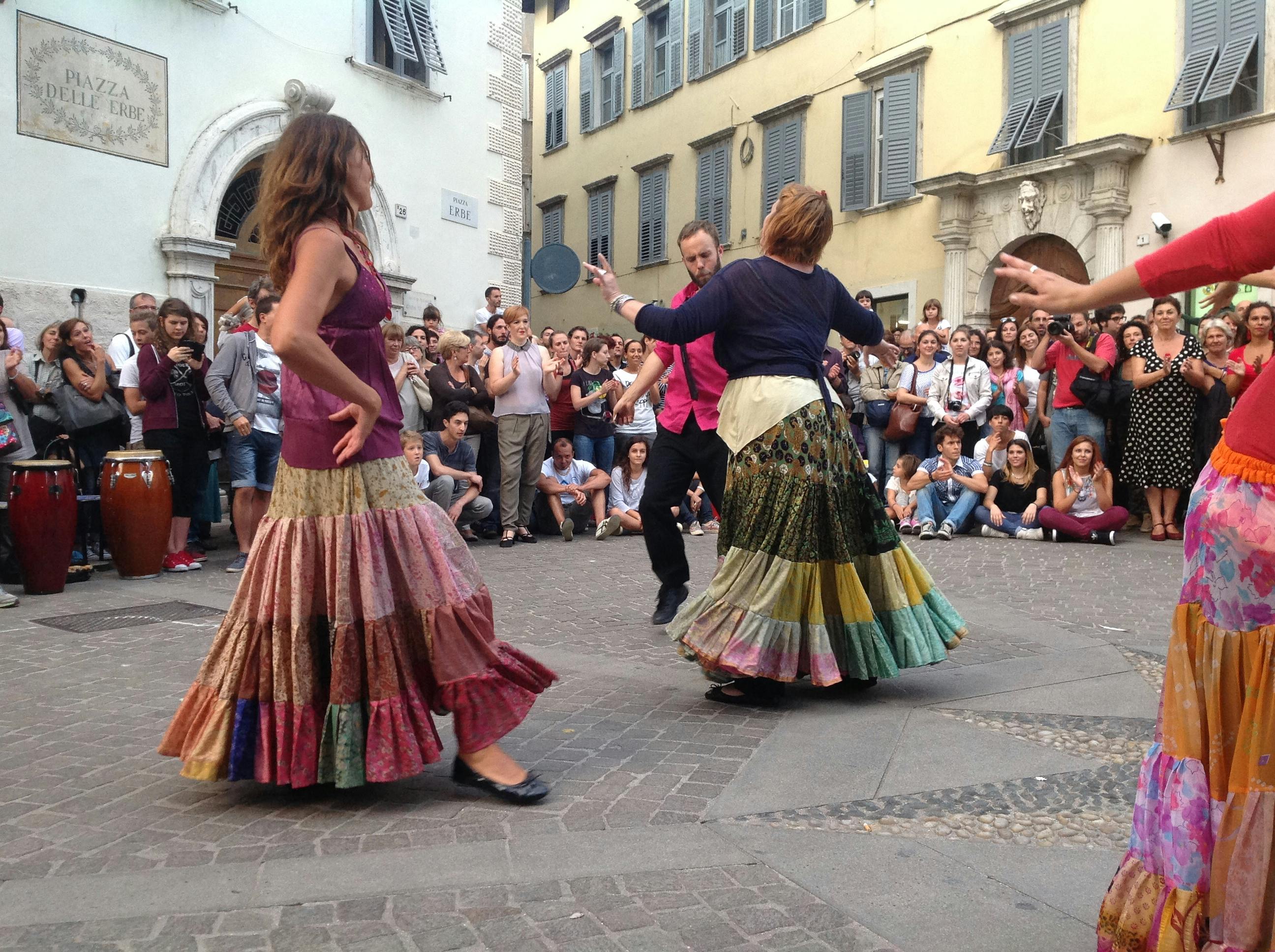 Couples are dancing in the center of Rovereto. The ballerinas are wearing long, colorful skirts. A large audience surrounds them.