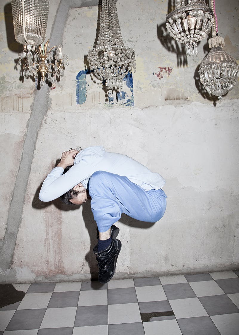 A man is in the air, curled up in a jump. He is wearing blue pants and a white shirt, hiding his head between his arms. Above him are small chandeliers.
