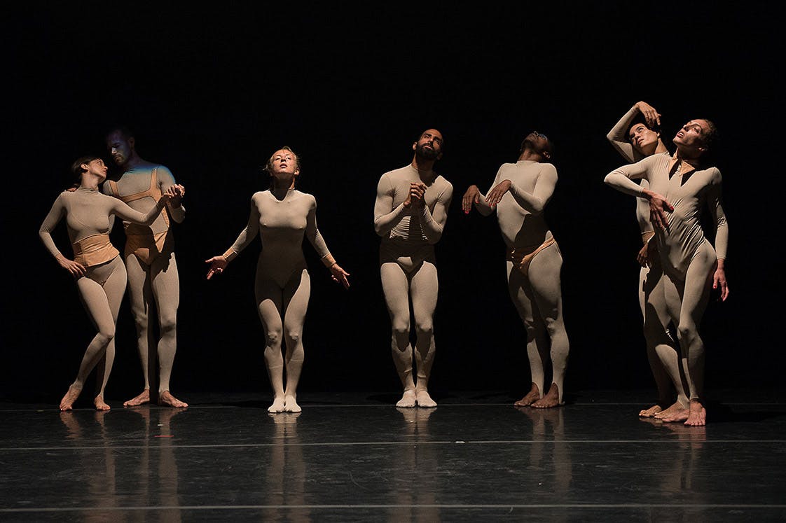 Seven dancers are on stage, standing in different poses. They wear close-fitting grey jumpsuits and are barefoot