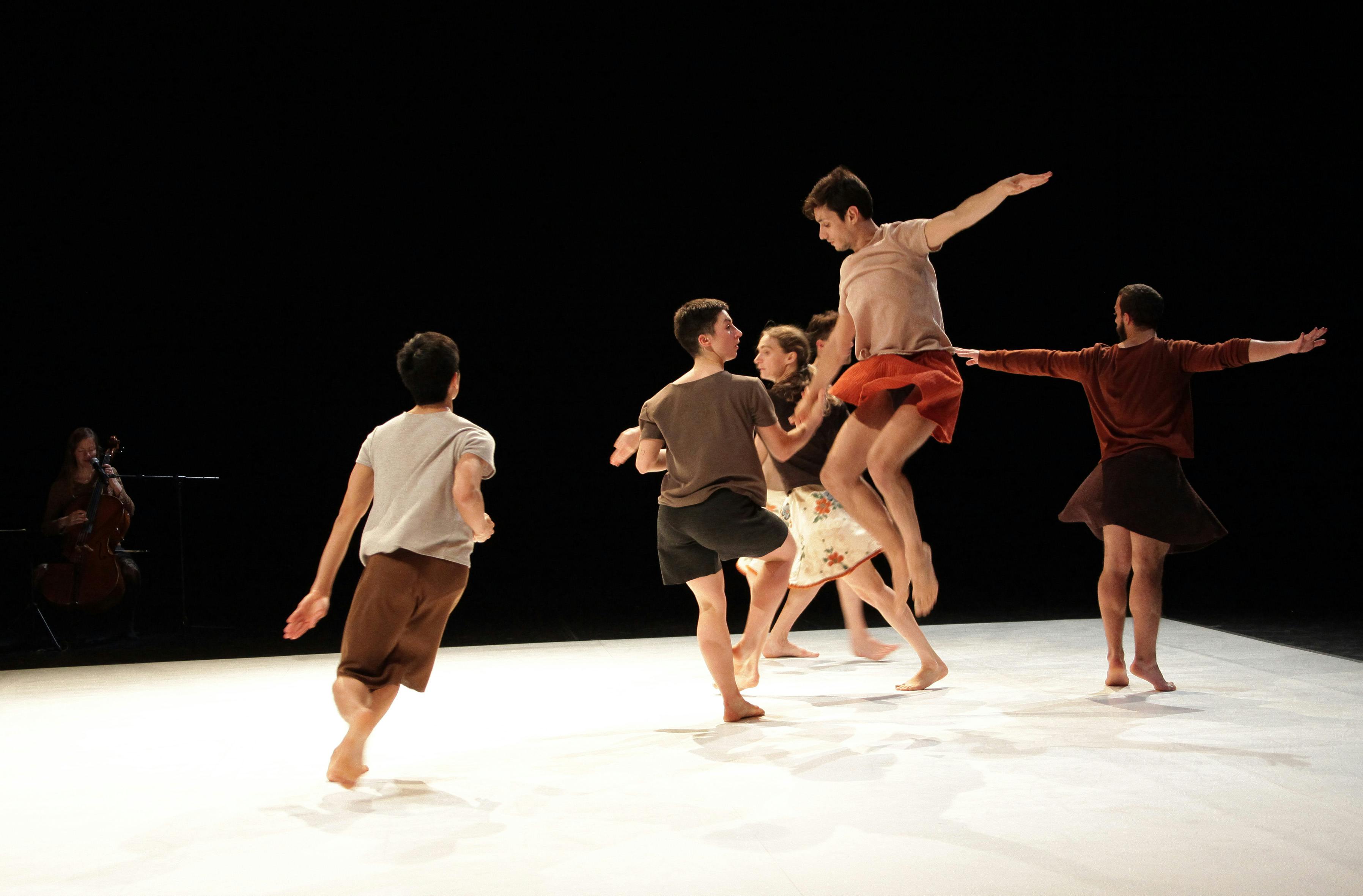 Six dancers are on stage, on a white carpet. Some are running while others are jumping in the air.