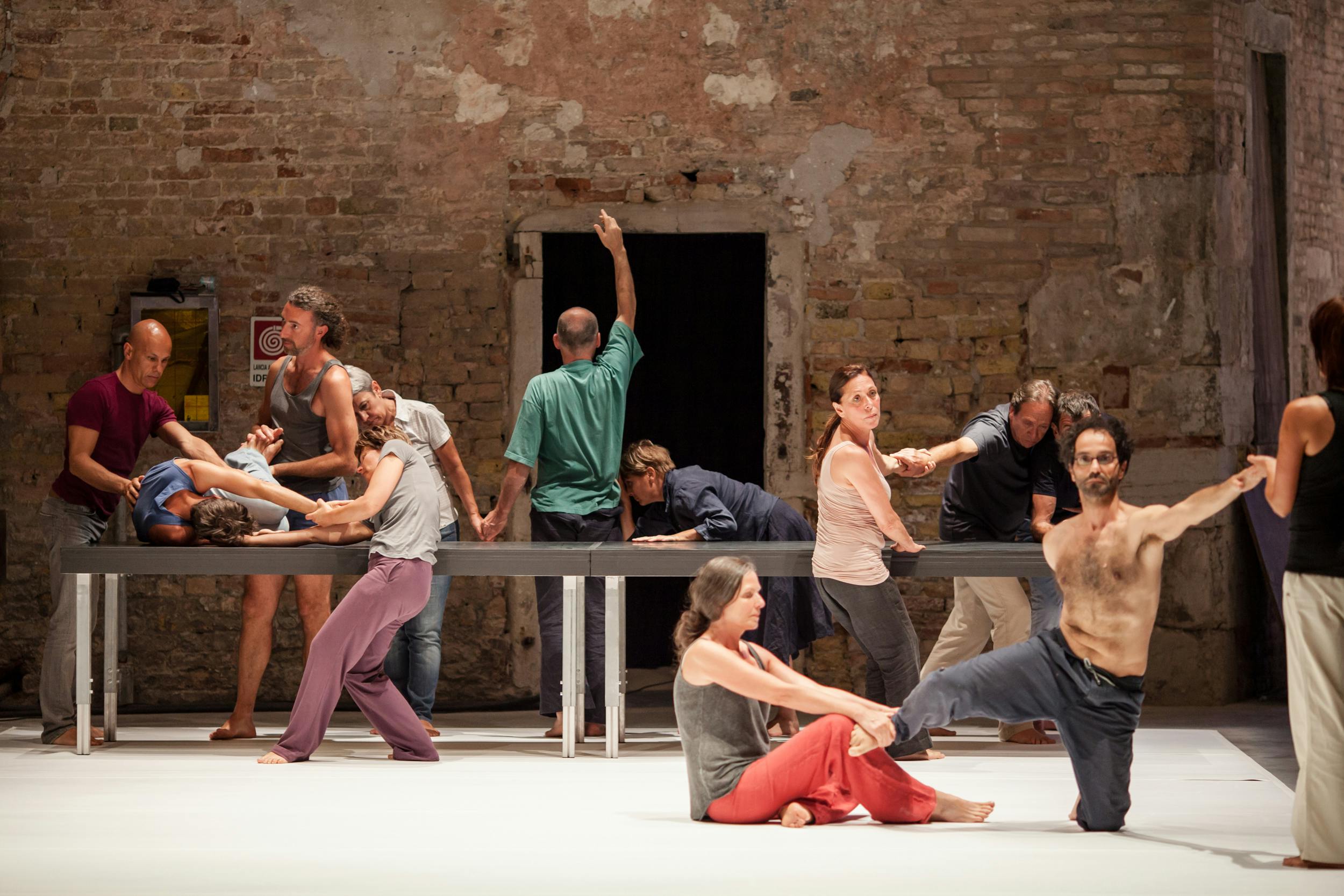 A dozen artists in various poses. There is also a very long table on stage. The background features a brick wall with a door in the middle.