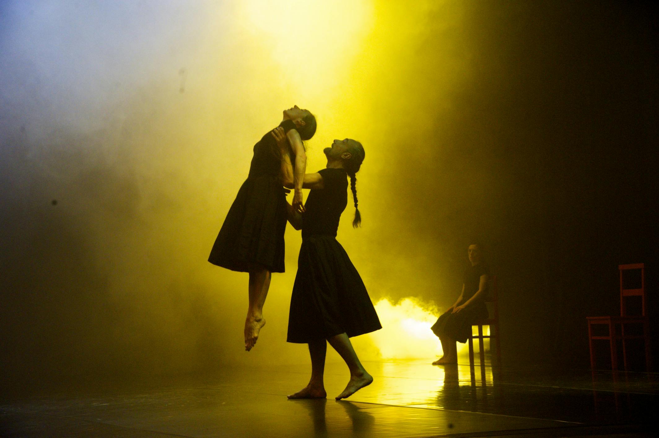 Luci gialle, tre artisti in scena. Uno tiene sospesa una ballerina in aria. Dietro di loro un'altra artista seduta su una sedia
