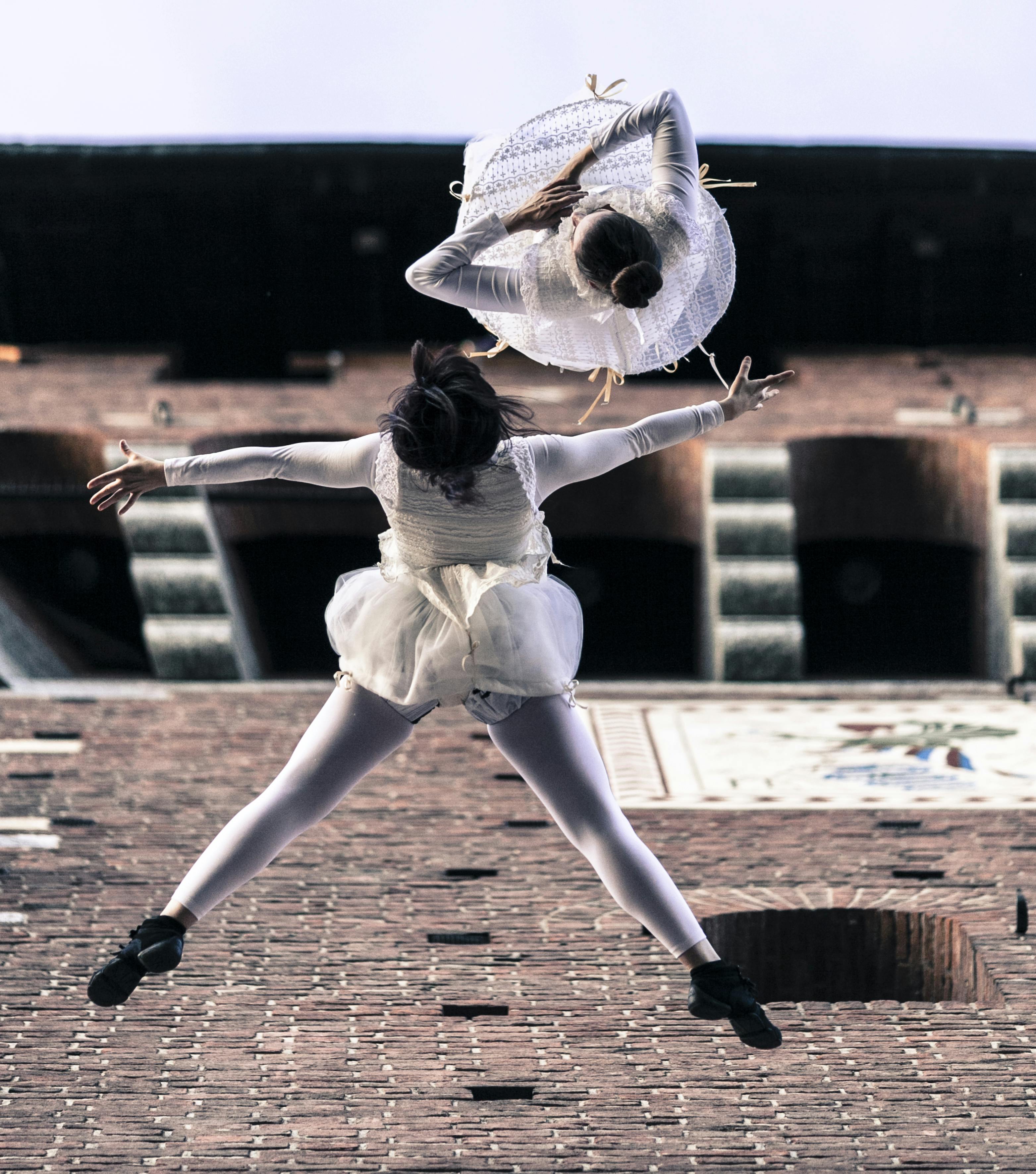 Two ballerinas in mid-air. They are dressed in white. One has wide arms and legs, while the other is upside down.