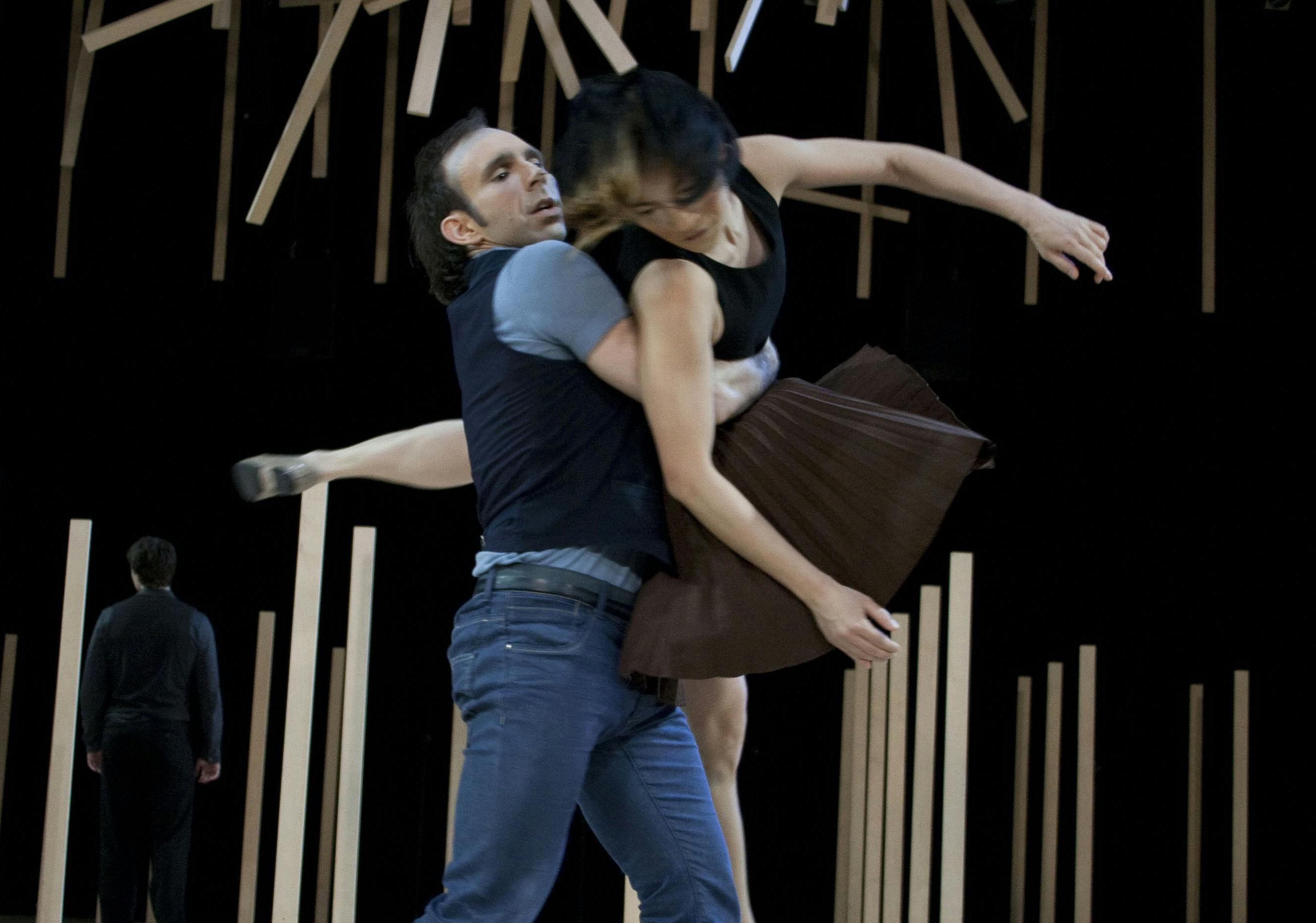 Due ballerini in scena, il ballerino sostiene la ballerina in braccio. Dietro di loro pali di legno verticali e sospesi