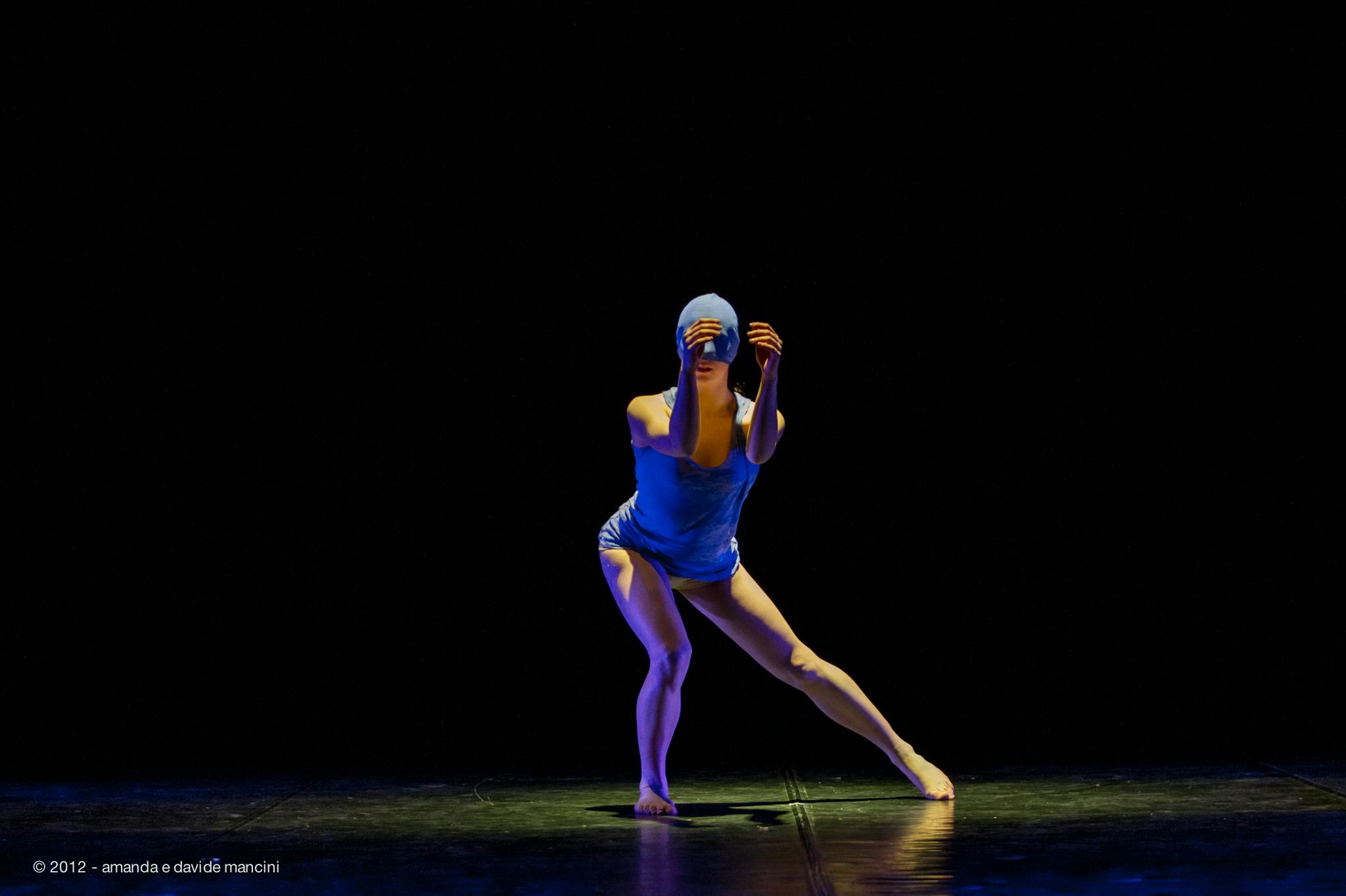 Una ballerina, in body blu, sul palco. è piegata su un ginocchio, ha le mani avanti a sè.