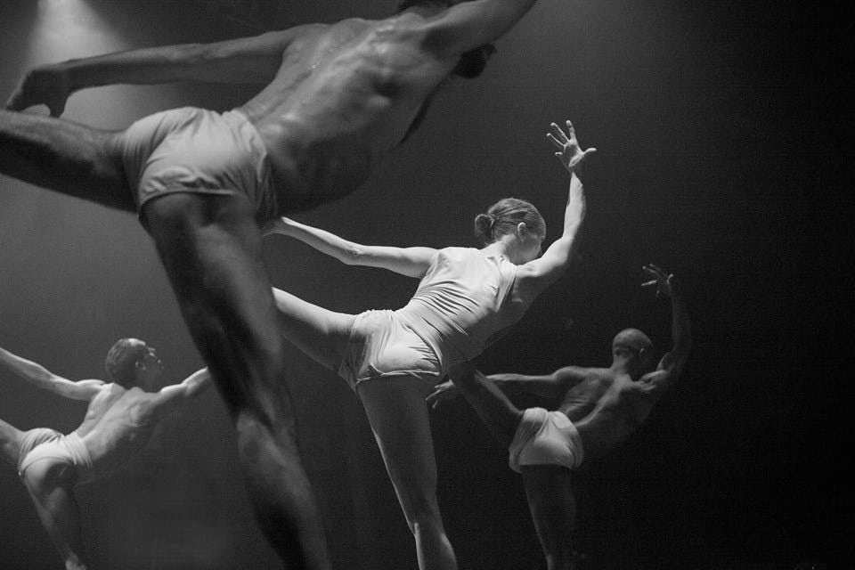 Black and white photo of four dancers. Their left leg is stretched backward in the air, and their right arm is extended upward.