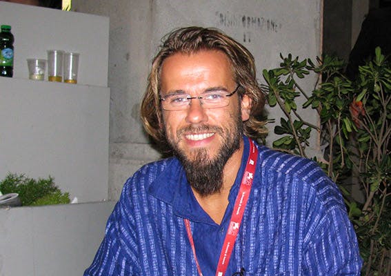 In foto Andrea Segre, regista di cinema documentario e di finzione. Sorride alla telecamera. Indossa occhiali da vista e una maglia blu.