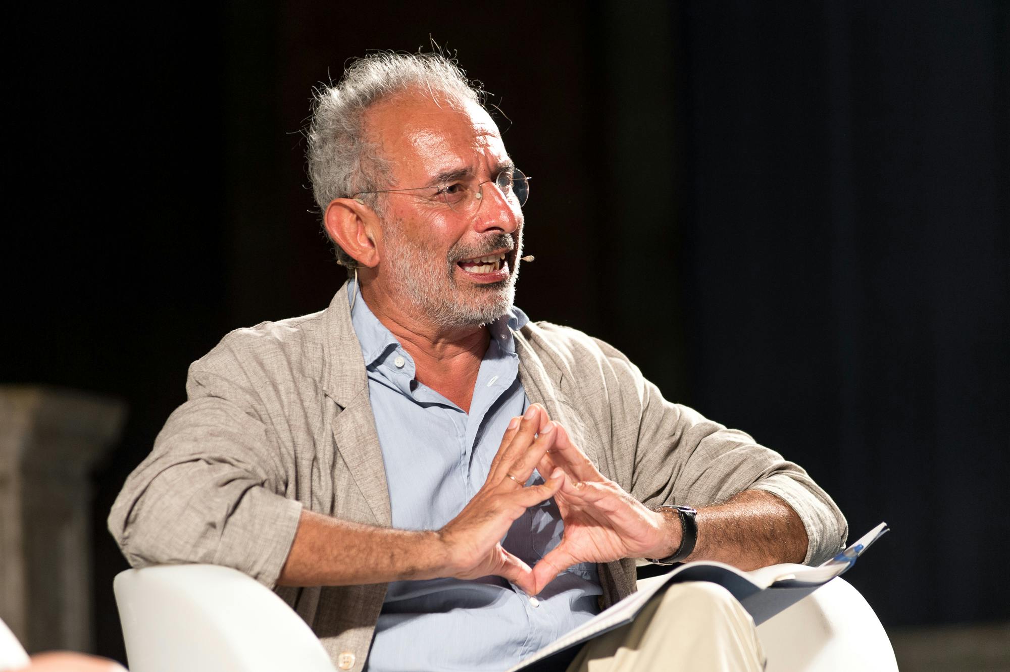 In the photo, Gad Lerner, journalist, television host, and essayist, is sitting while speaking. He has his hands clasped together and is wearing glasses.