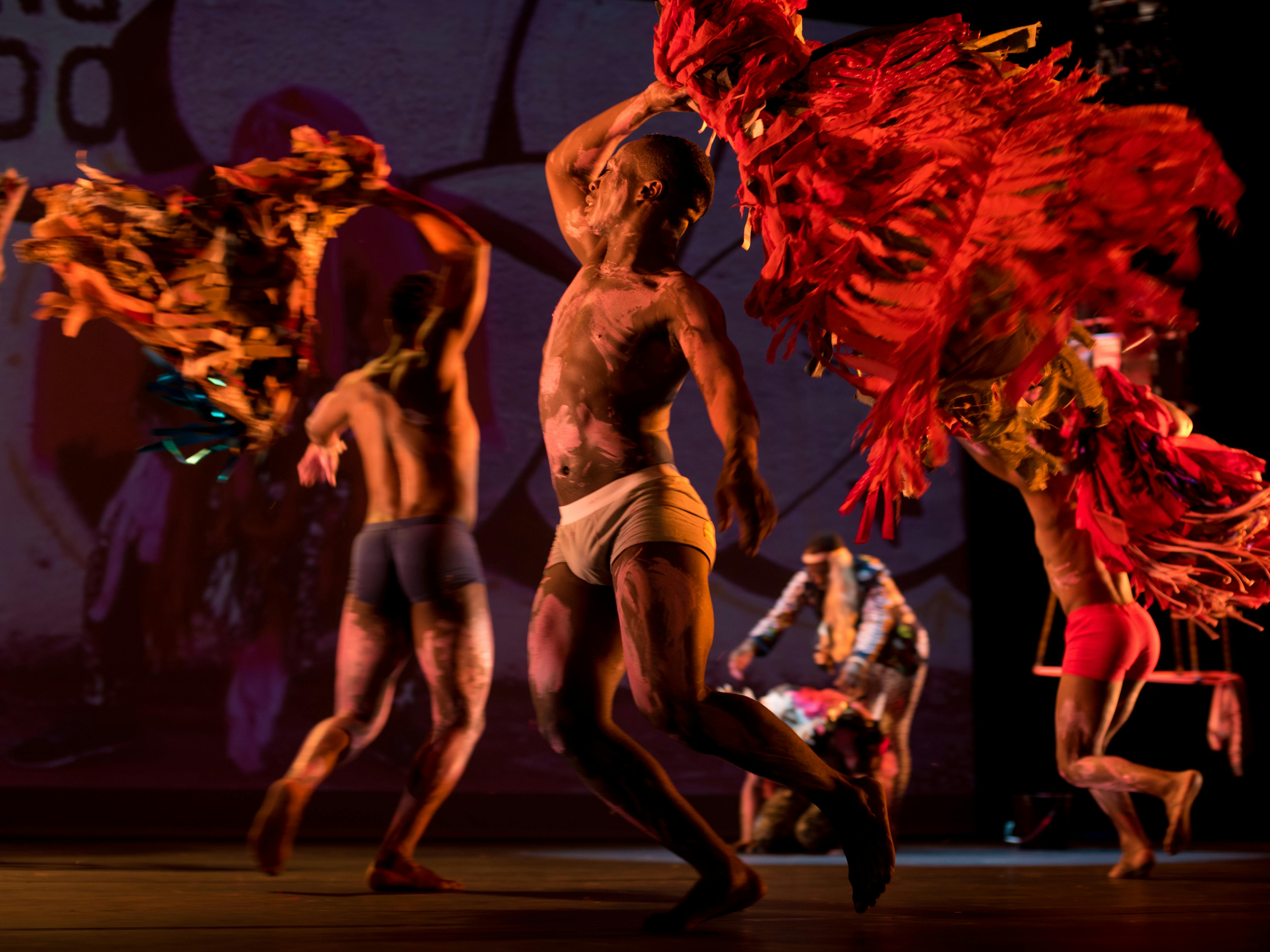 Three artists on stage, in an intimate setting. They are running in a circle, holding large feathered objects in their hands.