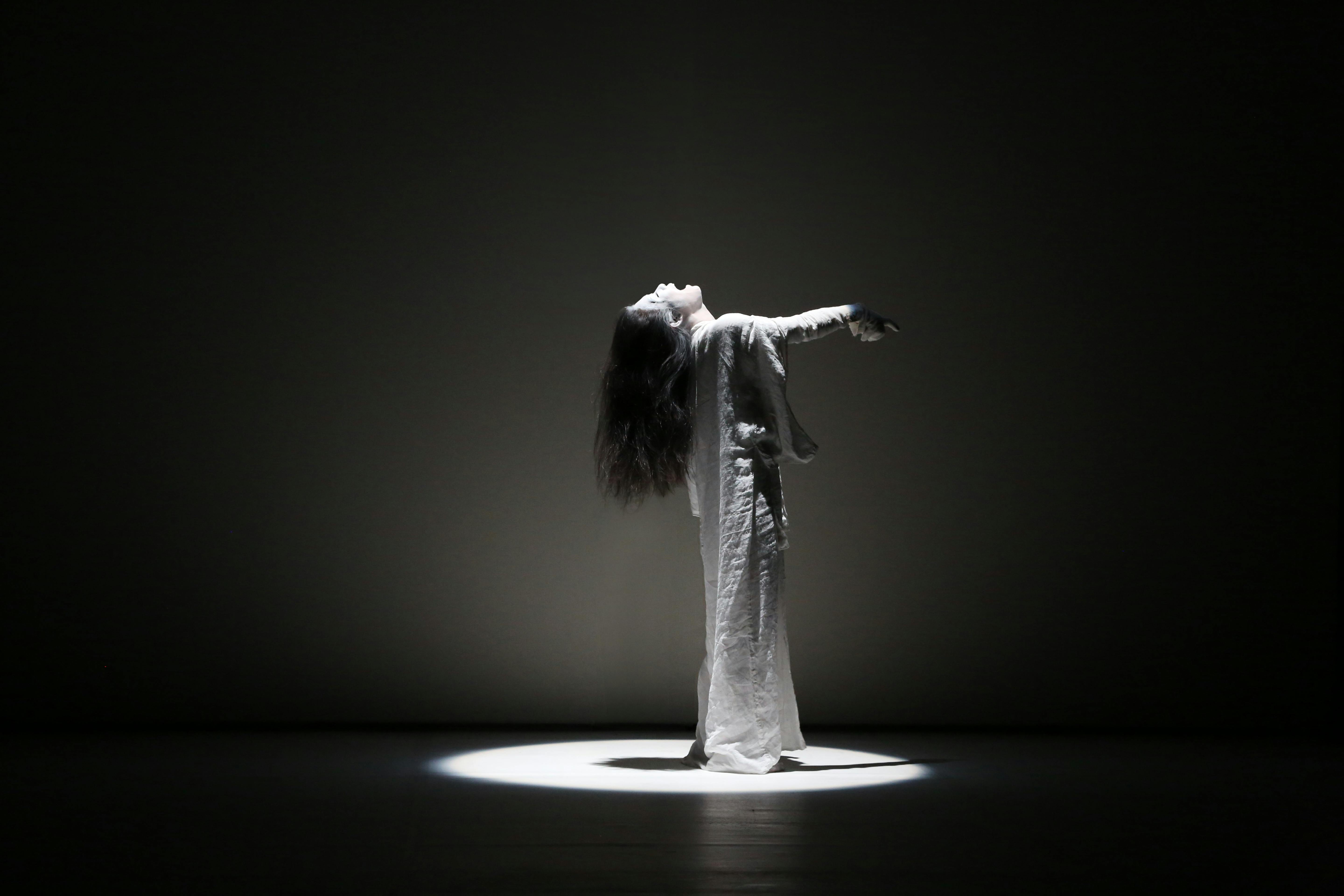 Black and white photo of an artist. She is standing and looking up. Her long hair falls backward. The figure is illuminated by a spotlight from above.