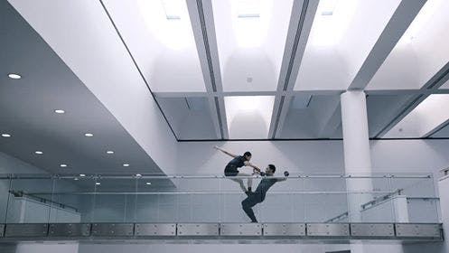 Foto di due artisti dietro a una balconata trasparente del MART. L'artista è in piedi sulle ginocchia dell'altro artista. Si tengono in equilibrio tenendosi per mano