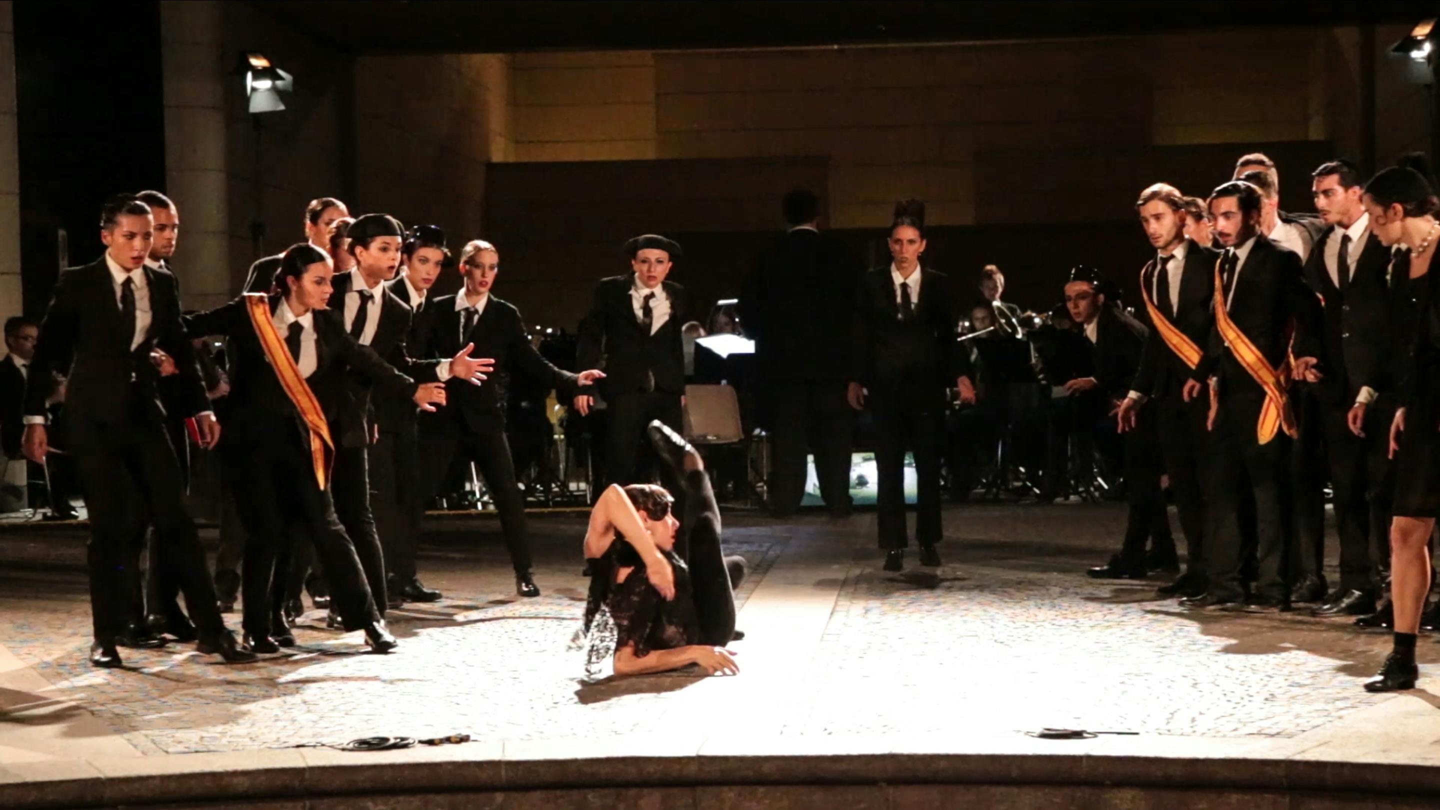 All'interno della piazza del Mart una artista è sdraiata a terra, in posa, con la gamba alzata. Intorno a lei una ventina di artisti in cerchio, la guardano. Sono tutti vestiti di nero.