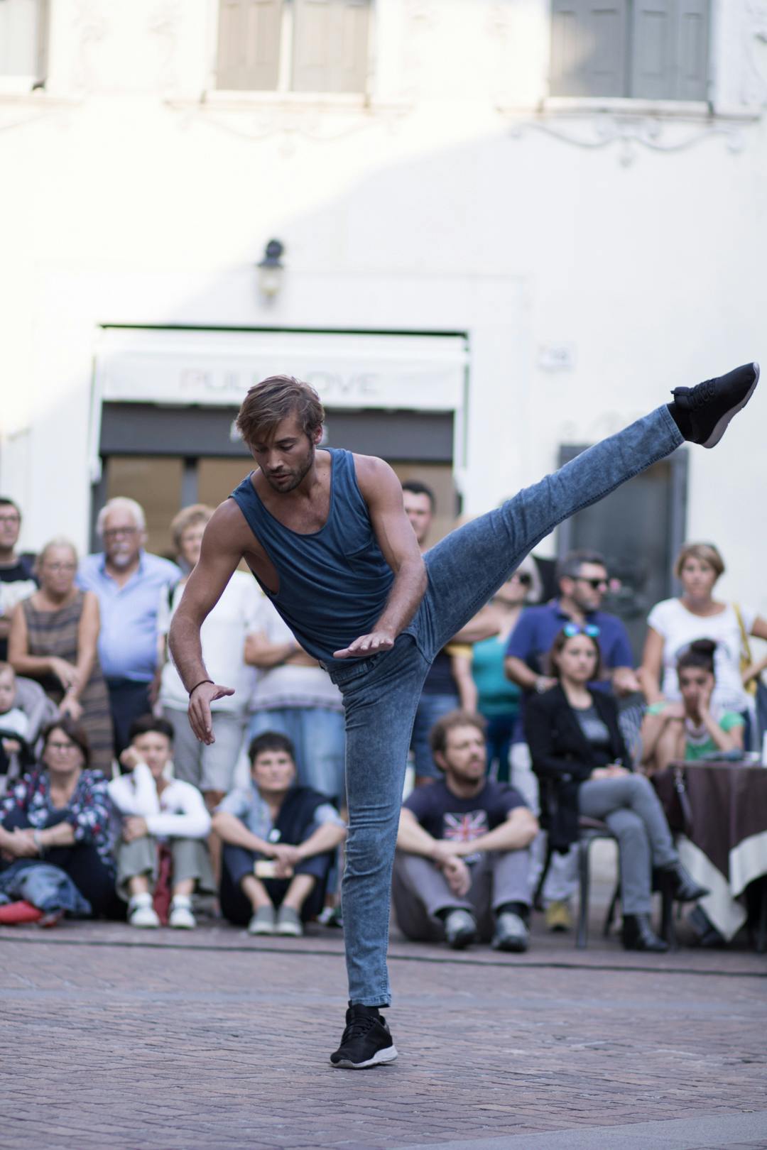 Un ballerino in mezzo a una piazza. Ha pantaloni e canotta azzurri. Ha la propria gamba sinistra tesi in aria. Dietro di lui pubblico seduto a terra.