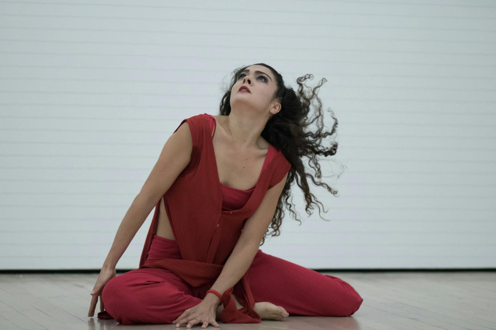 An artist on stage is sitting on the ground. She is dressed in red and has long black hair. She is looking up.