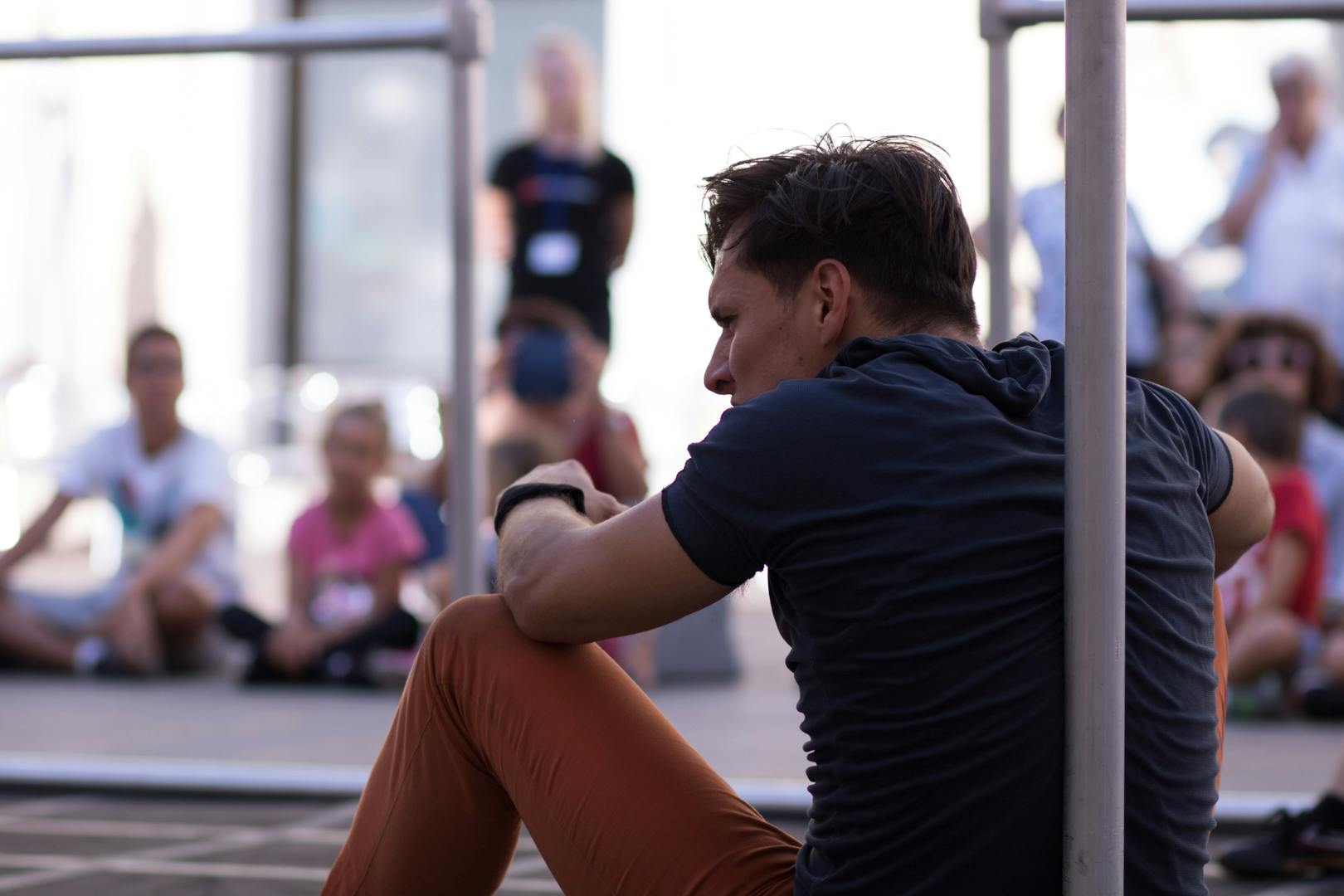 Photo of an artist from behind. He is seated, leaning against a metal pole. Behind him, the audience is seated on the ground.