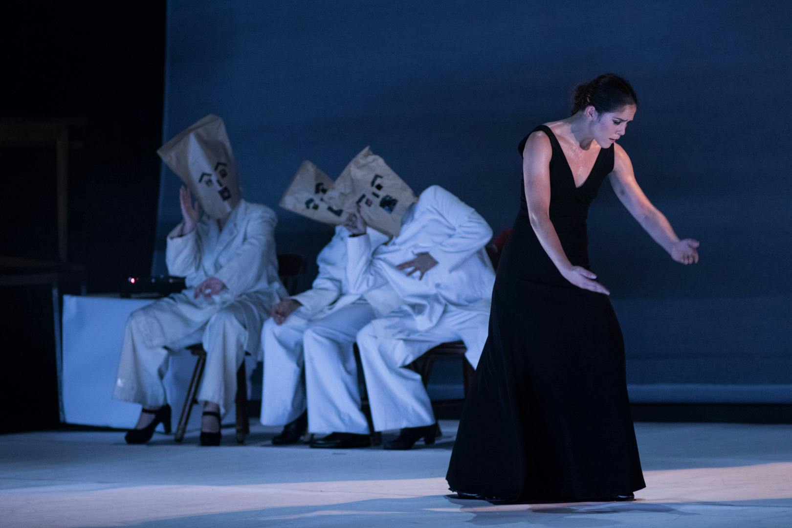Quattro artisti in scena. Una donna, con un vestito elegante nero è in piedi, guarda in basso. Dietro di lei tre artisti seduti: indossano tute bianche e dei sacchetti di carta sulla testa con delle facce disegnate a pennarello.