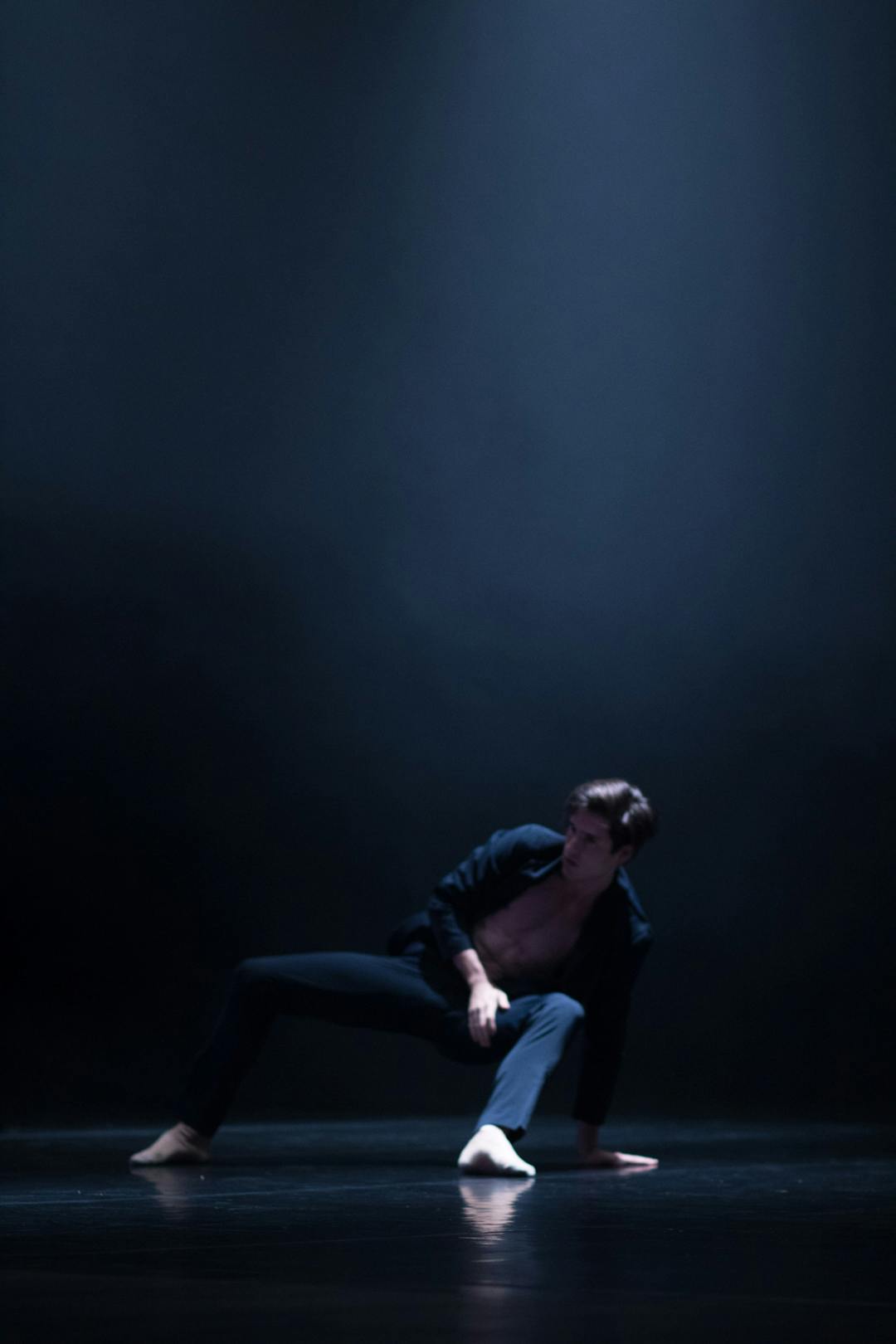 foto con luci molto scure. Al centro un ballerino, vestito di nero. Ha entrambi i piedi e una sola mano a terra