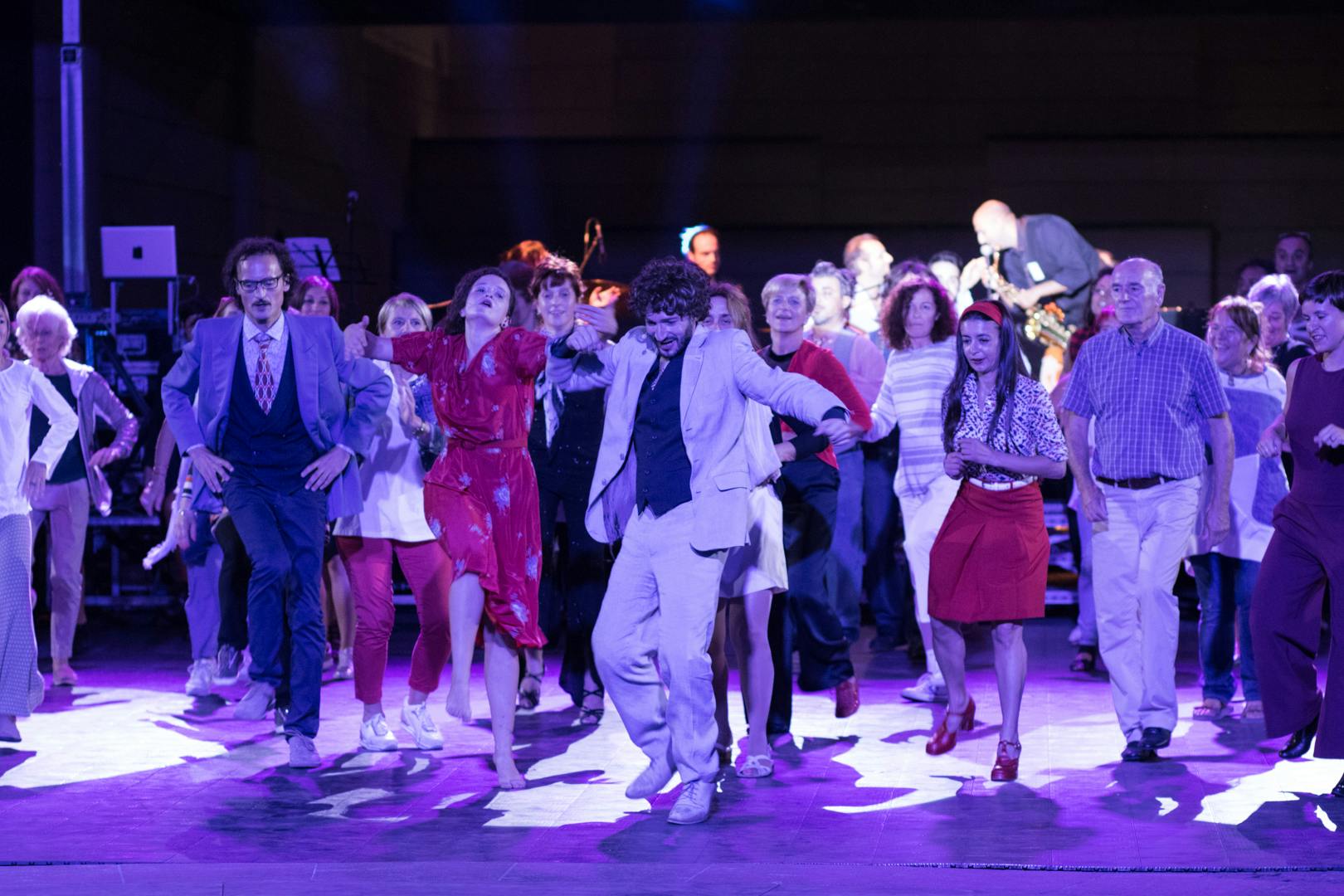 Many people on the dance floor, some artists, some from the audience. They are standing and dancing next to each other. In front, an artist in a white outfit