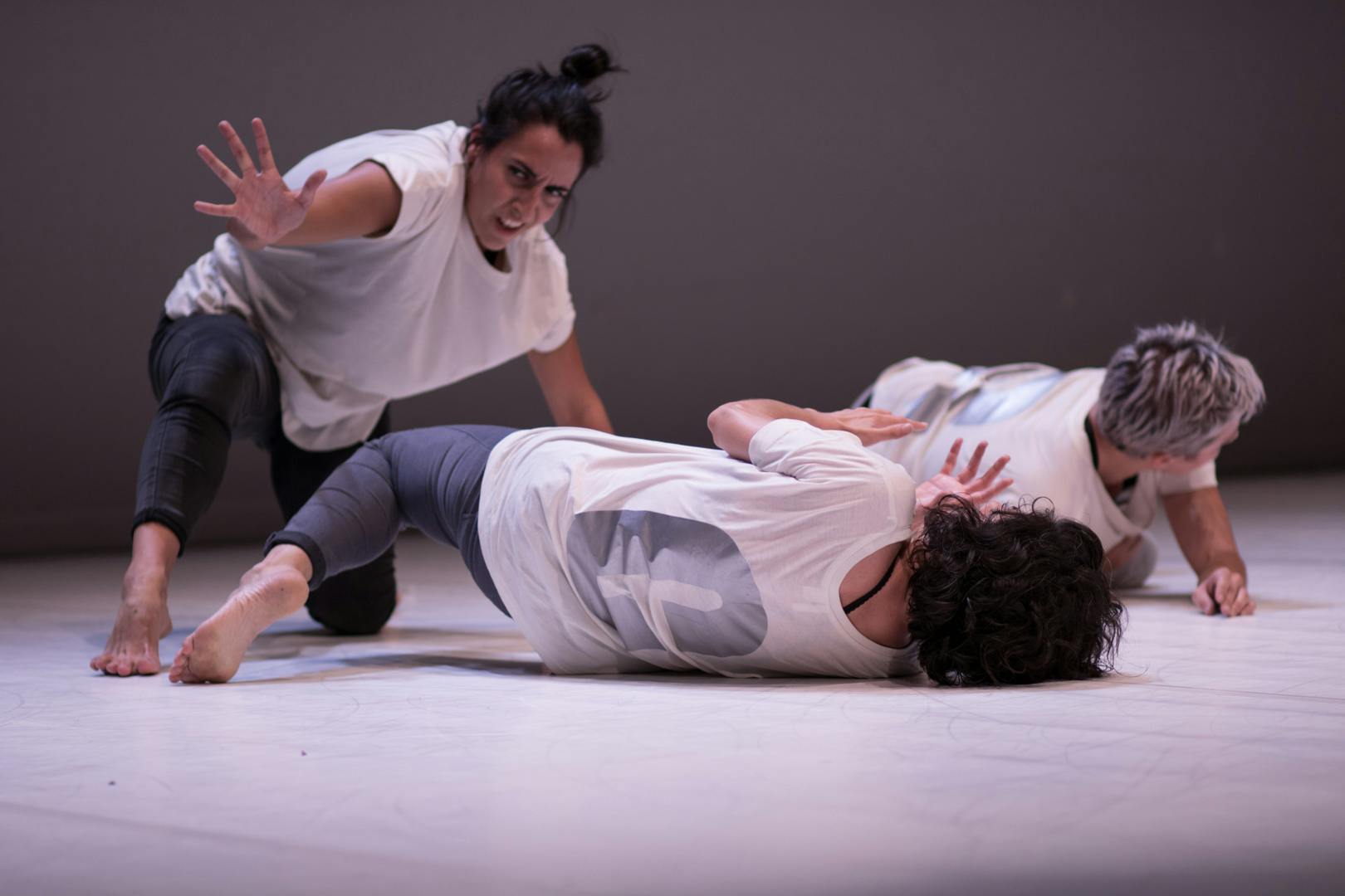 Three performers on stage, wearing dark pants and loose white shirts. Two are lying on the ground in motion, while the other is leaning on her knee.