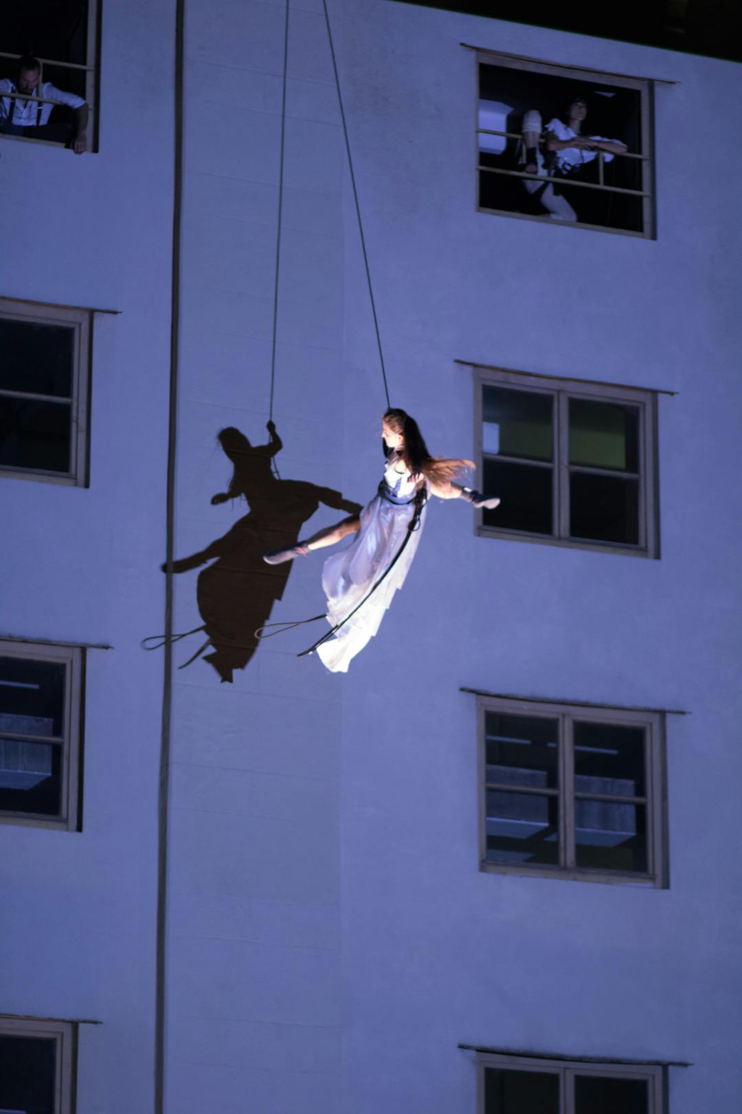 Photo of an artist in flight, attached with a harness. Her body casts a shadow on a building