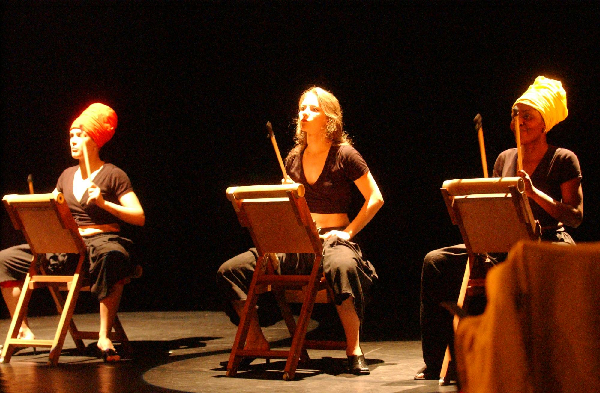Tre musiciste sul palco, sedute. Tengono in mano delle bacchette da tamburo e davanti a loro hanno degli oggetti quadrati.
