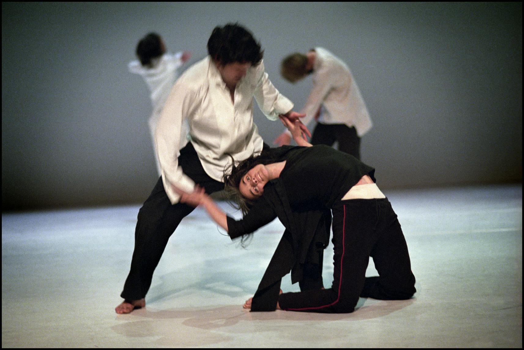 A dancer dressed in black is kneeling on the ground, arching her back backwards while a standing male dancer holds both her hands. In the background, two other artists are present.