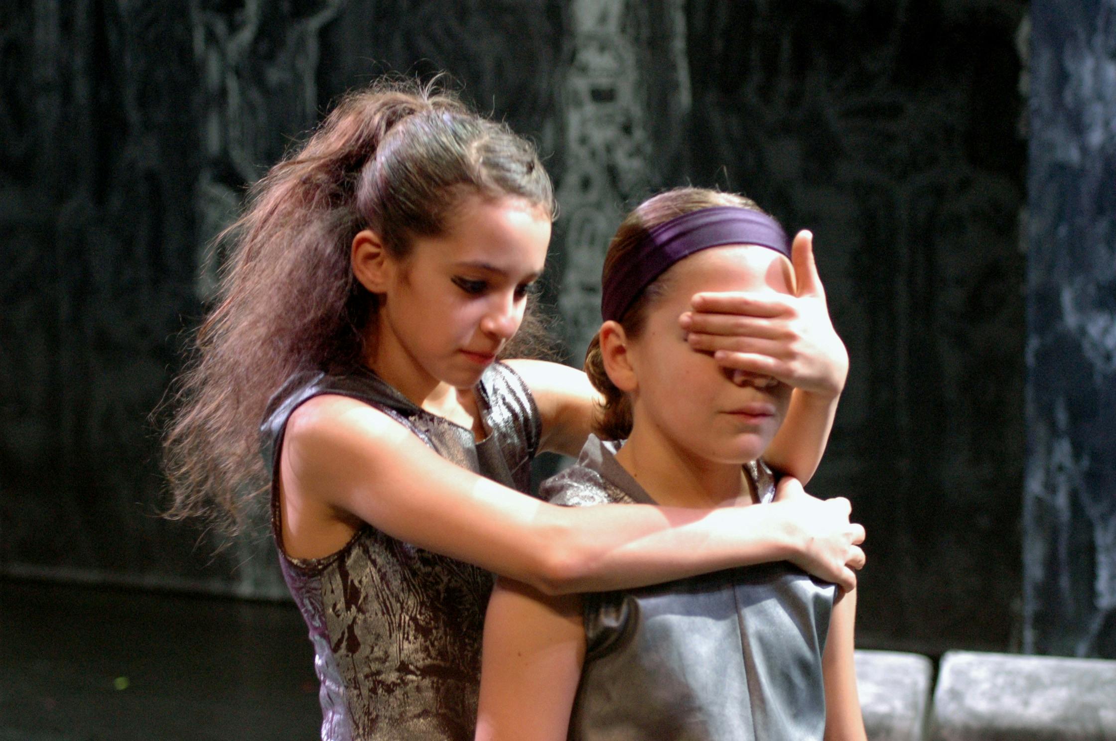 Due bambine in scena, uno dietro all'altra. Quella dietro copre gli occhi con la mano alla bambina davanti a lei e le tiene il braccio intorno alle spalle.