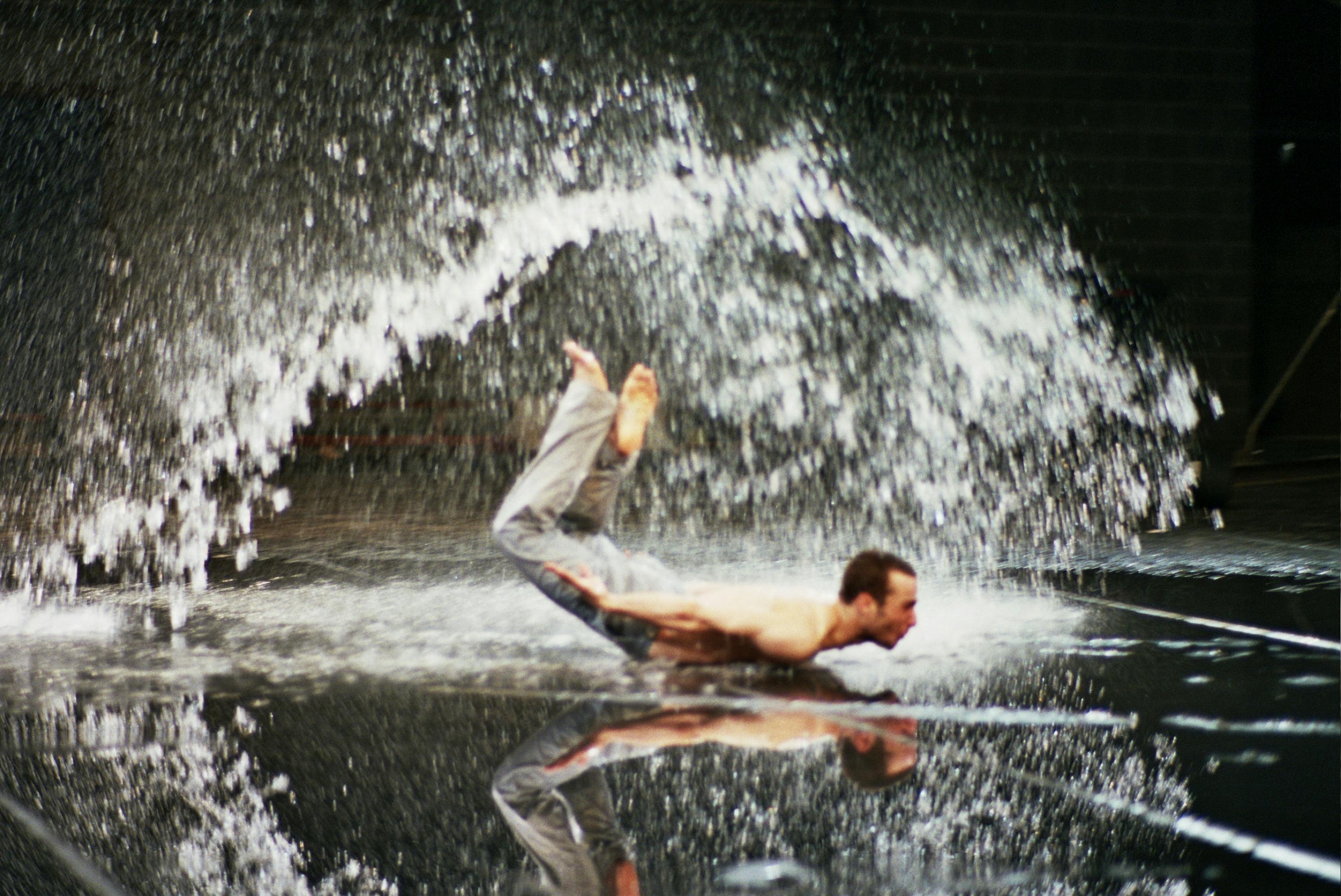 A shirtless dancer lies prone on the floor, with arms and legs slightly lifted. Around him, splashes of water.