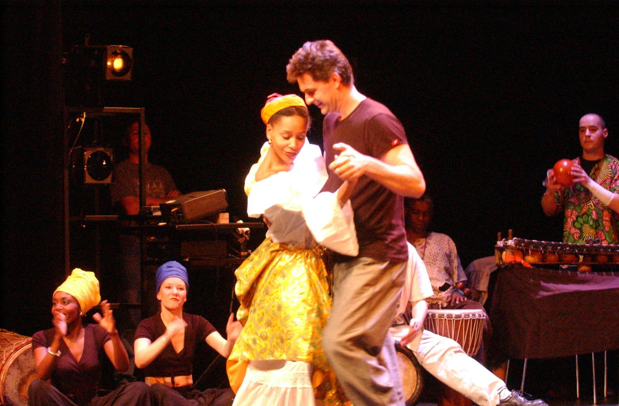 Two dancers are dancing in the center of the dance floor, smiling. Behind them, seated ballerinas are applauding and musicians.