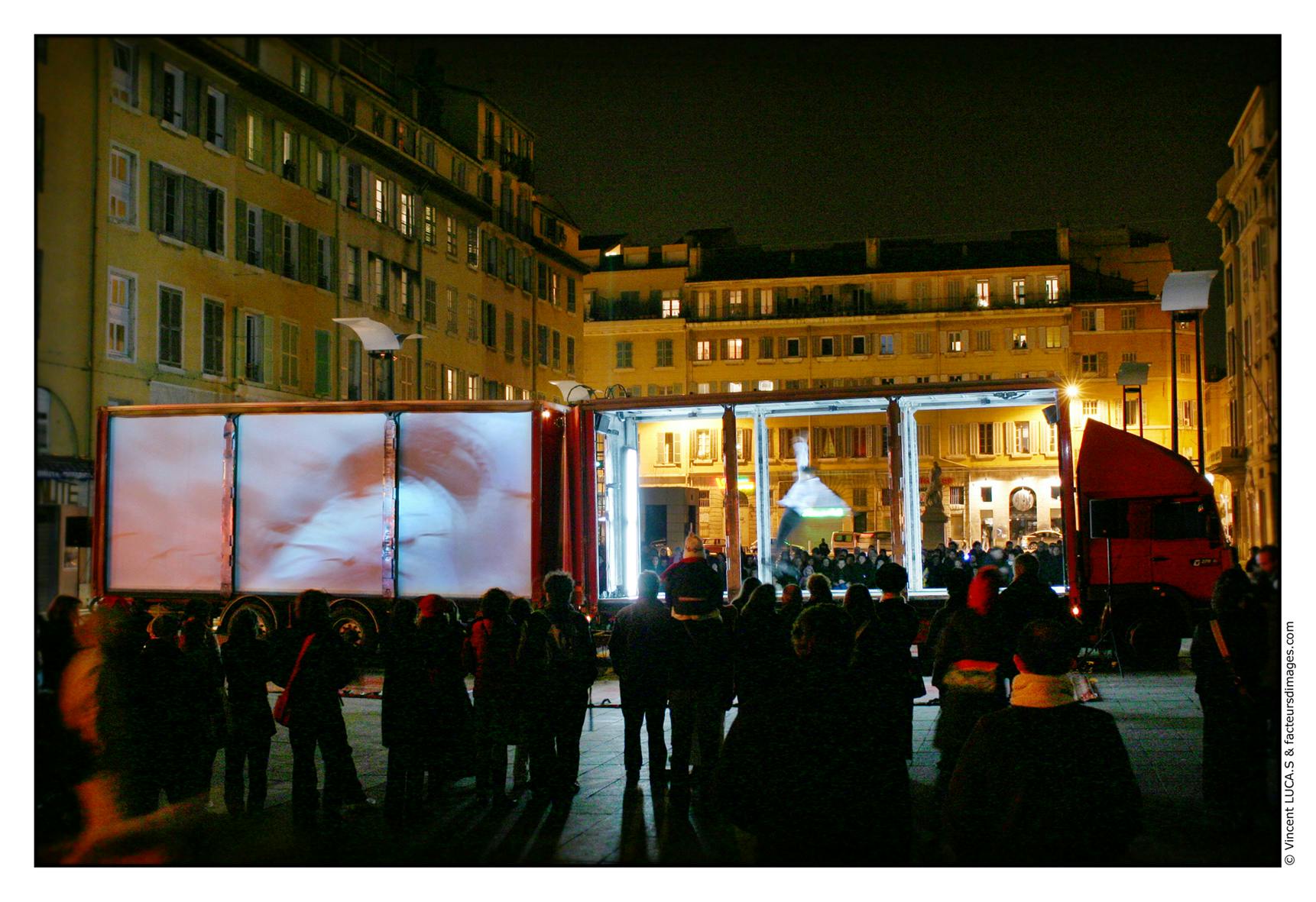 A crowd watching a performance in a square with a truck without walls serving as the stage.