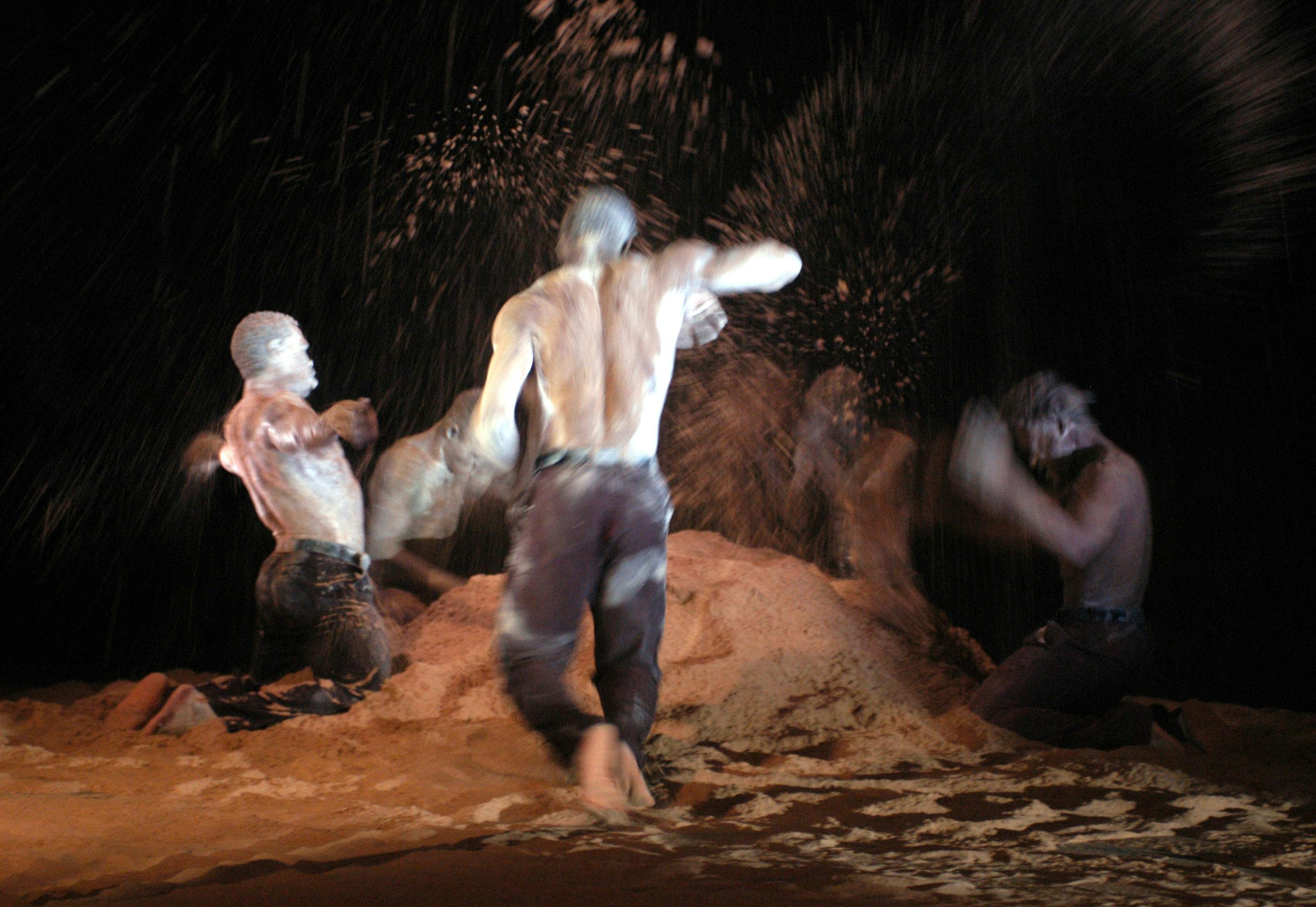 five artists bare-chested with dark pants around a pile of sand, some standing, some kneeling, throwing it into the air