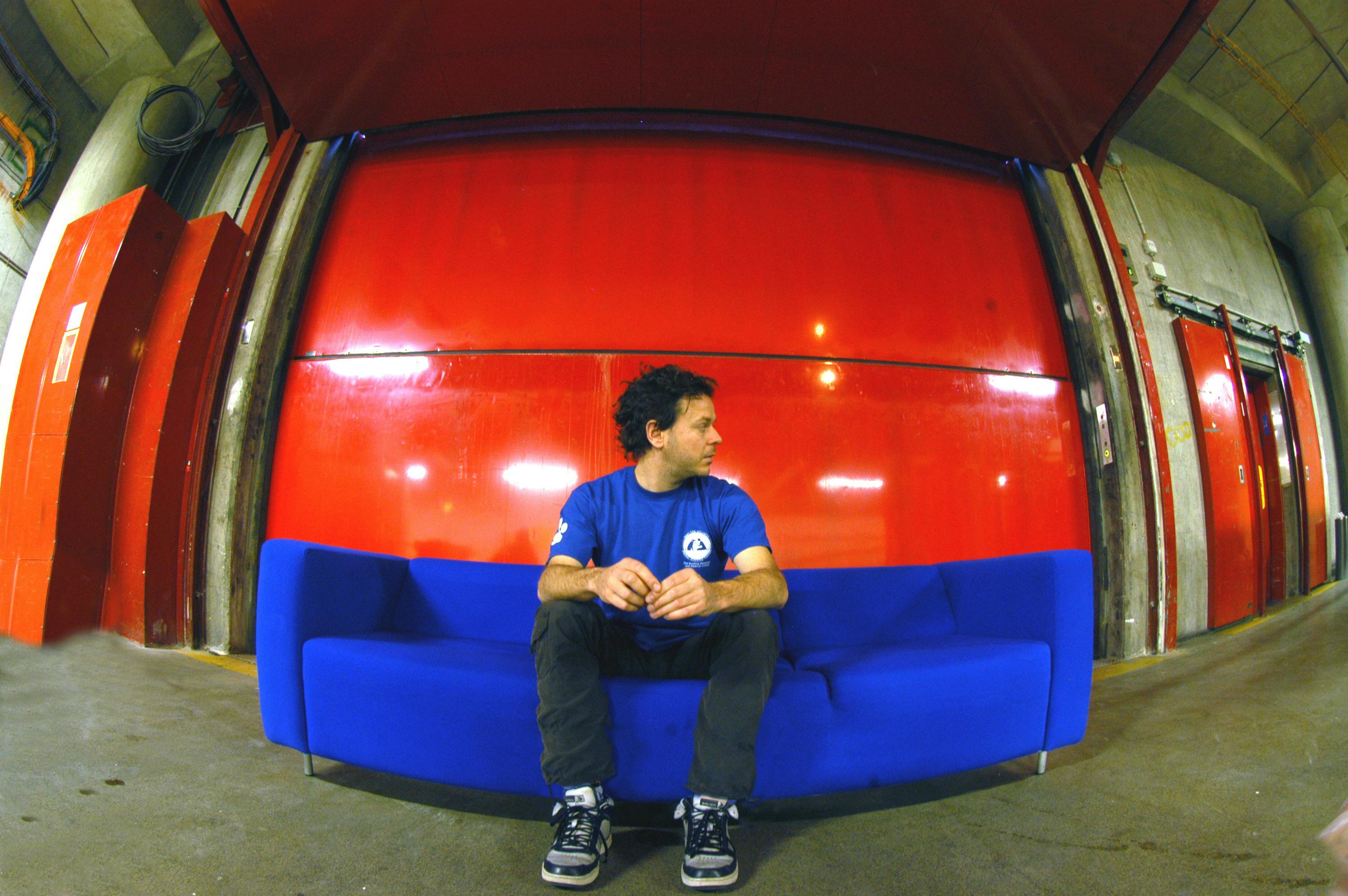 A person is portrayed sitting on a blue sofa, with a red background