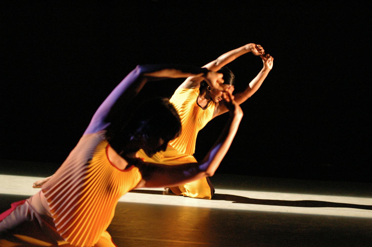 Two dancers, one in the background and one in the foreground, are seated and leaning their bodies to the right with their arms.