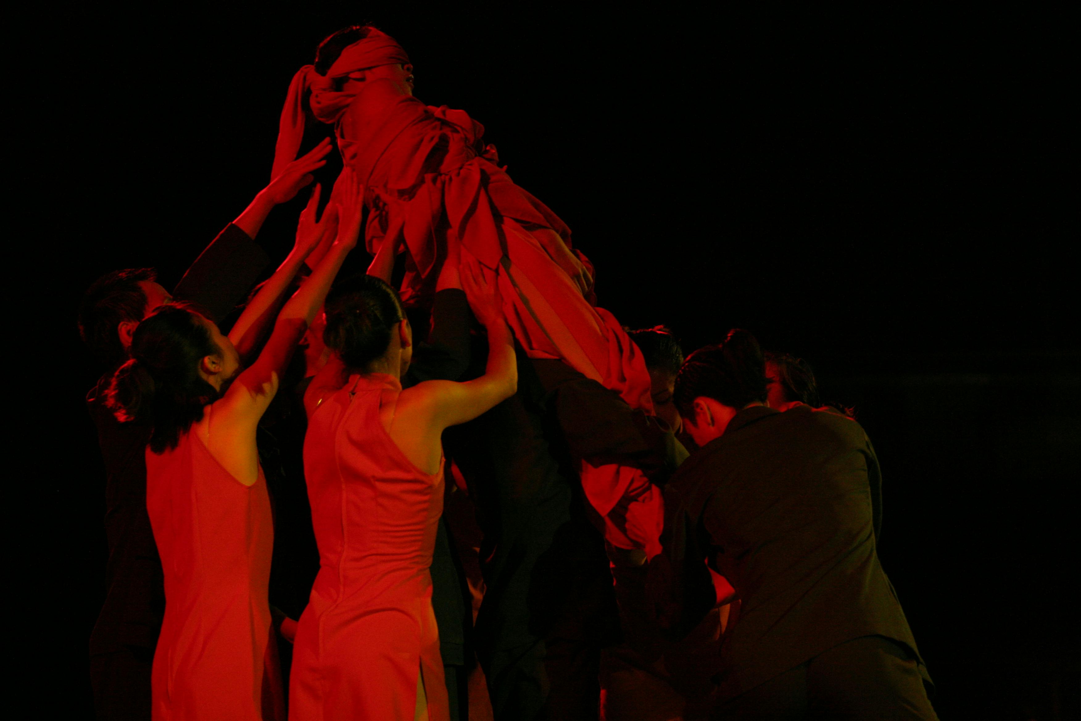con una luce rossa nella penombra dei ballerini stanno trasportando con le braccia in alto un altro artista avvolto in un telo