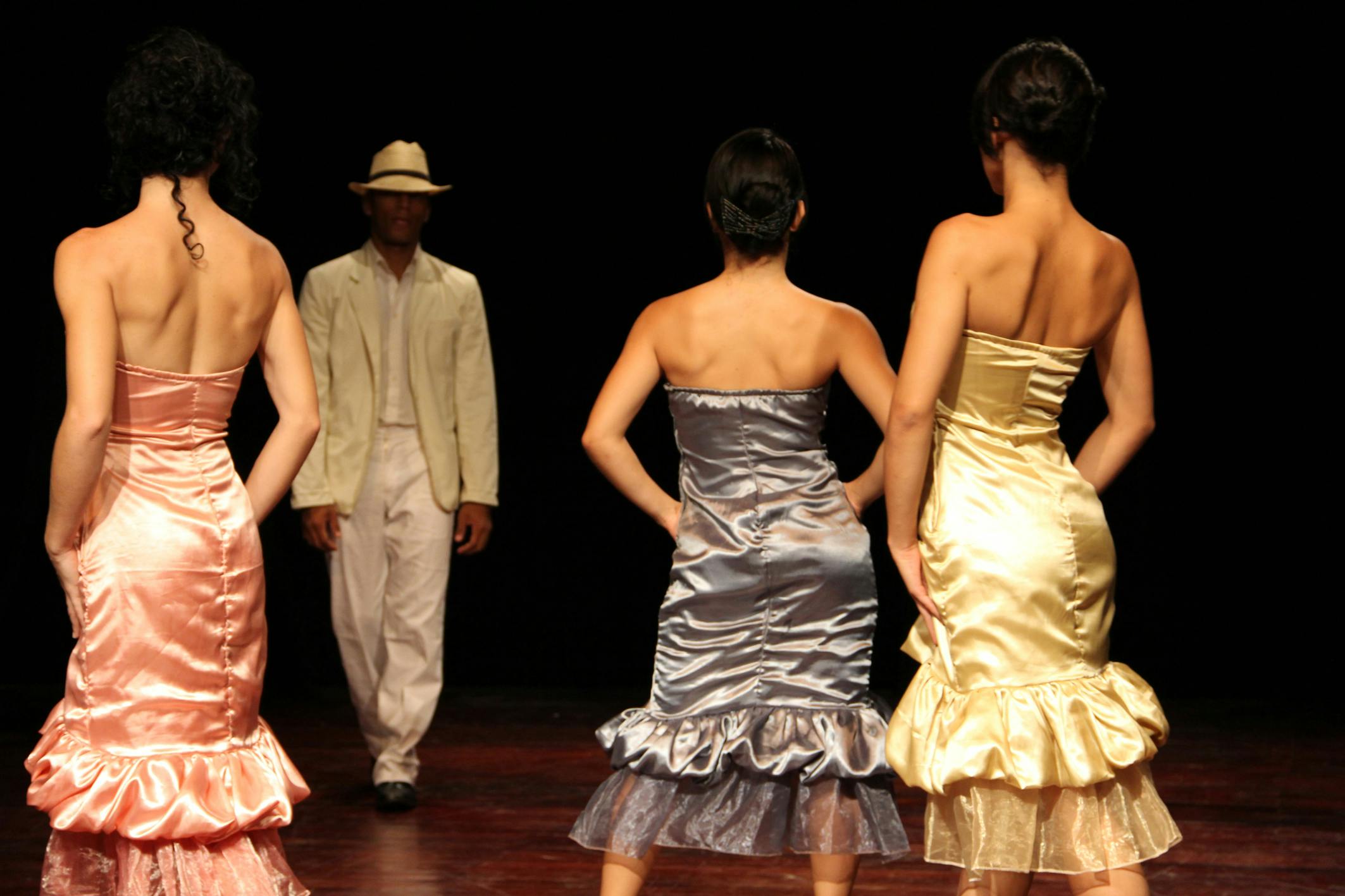 Tre ballerine di spalle, vestite con abiti colorati senza spalle. Guardano un uomo che cammina verso di loro.