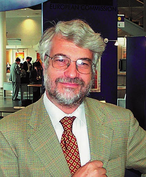 Portrait of Gianni Silvestrini. He has gray hair and a beard and wears glasses. He is dressed in a light green suit with a red tie.